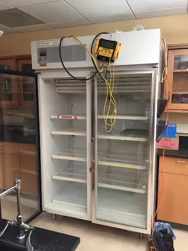 A large, white double-door refrigerator with an attached temperature monitoring device in a laboratory setting.