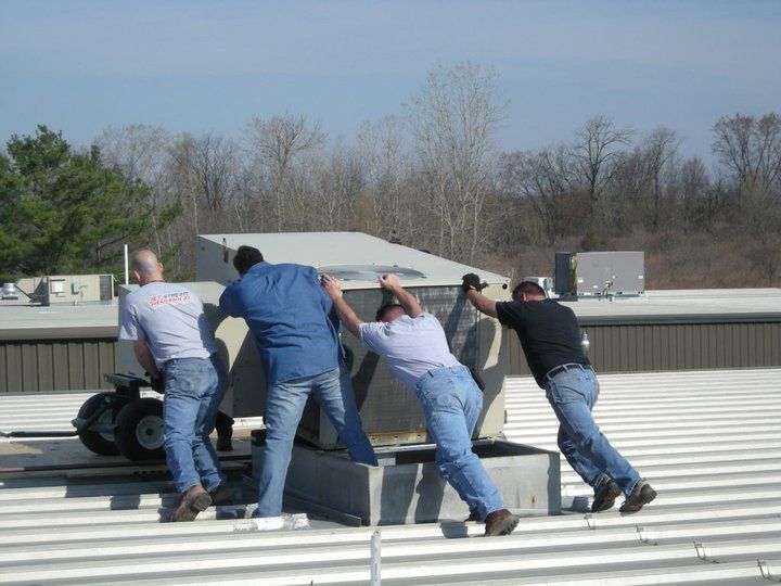 Four men in work clothes pushing an air conditioning unit on a rooftop.