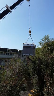 Crane lifting a large, grey industrial unit against a blue sky, over trees and a building.
