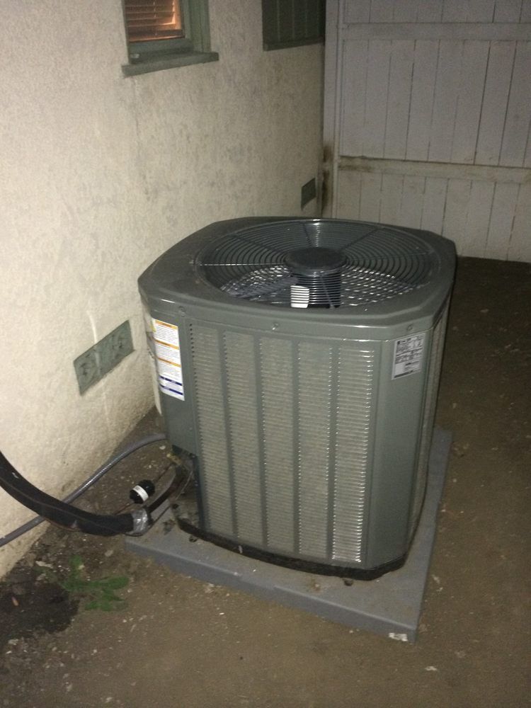 Gray air conditioning unit on a concrete pad near a wall and wooden fence.