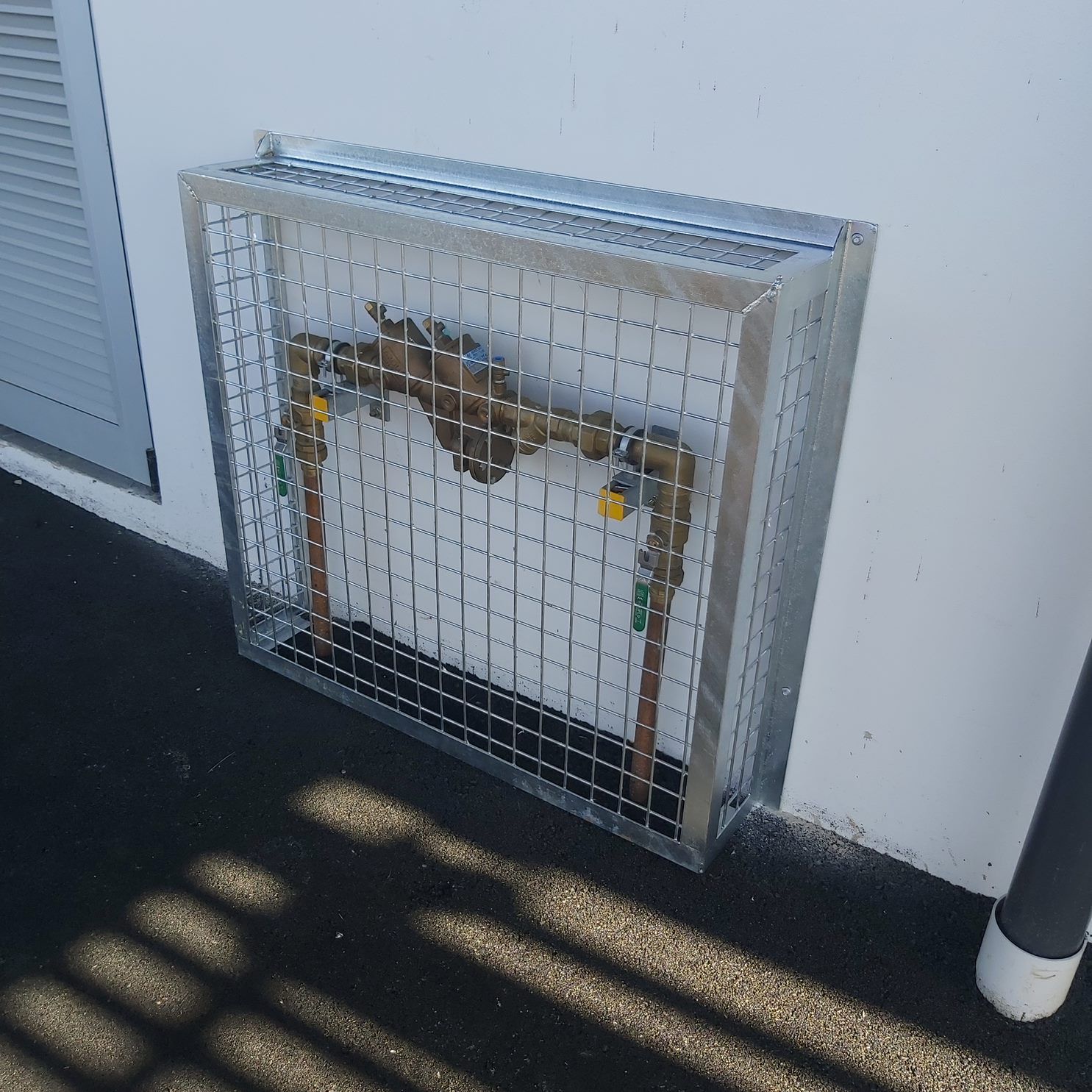 A caged metal enclosure protects water pipes and valves mounted on a white wall.