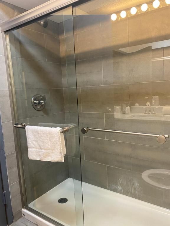 Shower stall with glass doors, white towel on the handle. Gray tiled walls, showerhead, and a vanity reflected in the glass.