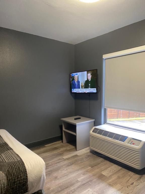 Hotel room: bed, TV on wall, nightstand, window with blinds, AC unit. Gray walls, light wood floor.