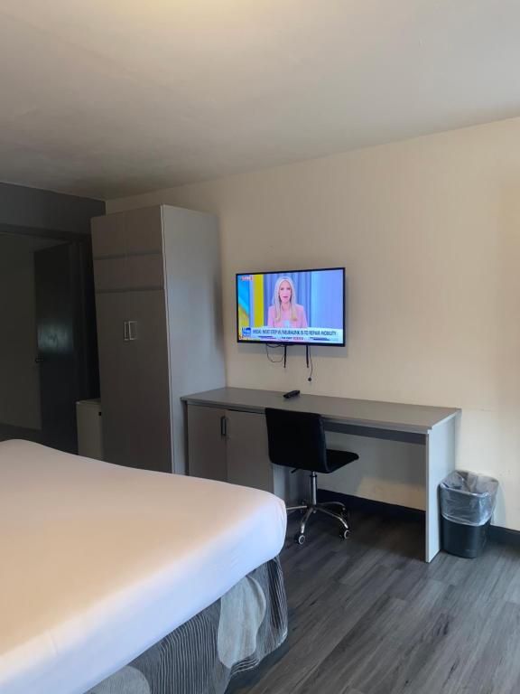 Hotel room with a bed, desk with chair, TV showing news, and a closet. Gray and white colors.