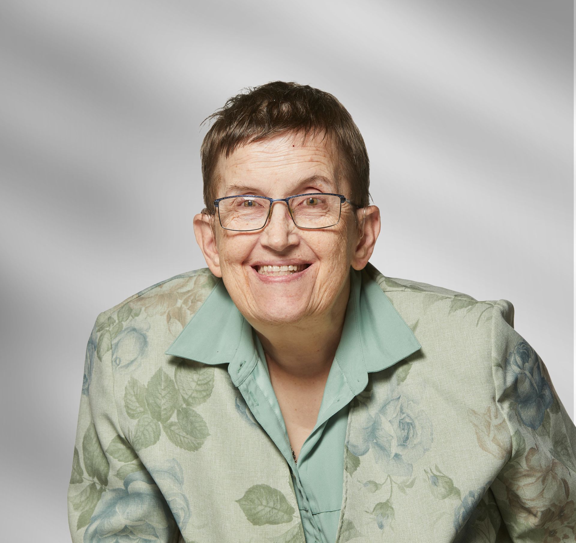 An elderly woman wearing glasses and a green jacket is smiling