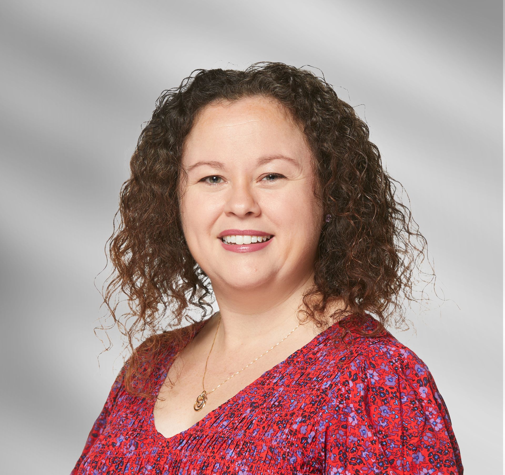 A woman with curly hair is wearing a red floral shirt and smiling.