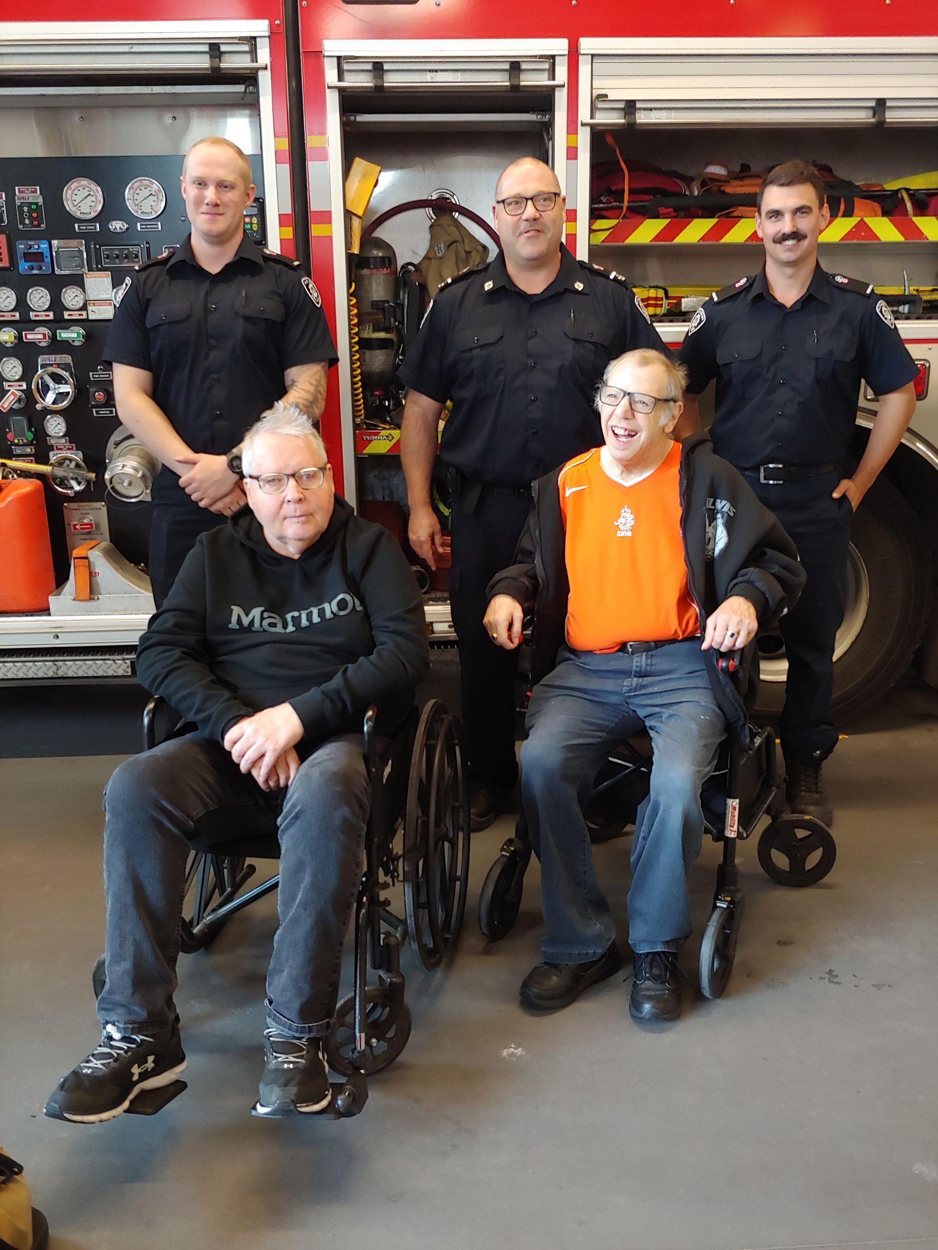 two men in wheelchairs infront of three firefighters with a fire truck in the background