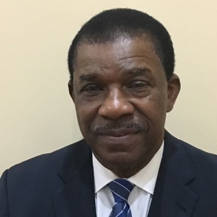 A Black man in a suit, with a mustache, smiling slightly; against a light tan background.