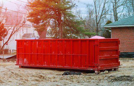 A large red dumpster is sitting in the dirt in front of a brick house.