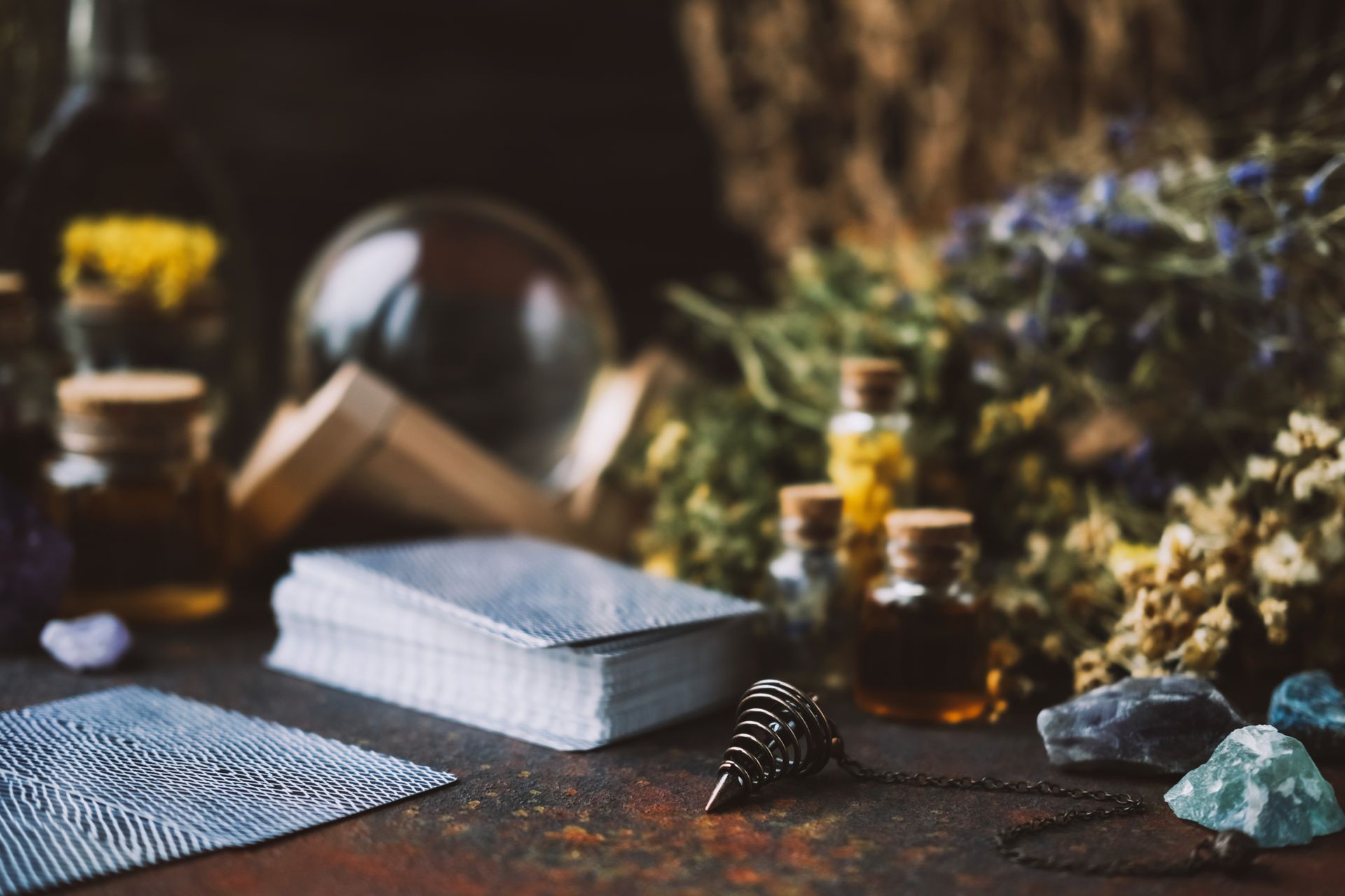 A pendulum, tarot cards, and stones on a table against blurred flowers, glass jars, & a crystal ball. A pendulum, tarot cards, and stones on a table against blurred flowers, glass jars, & a crystal ball.