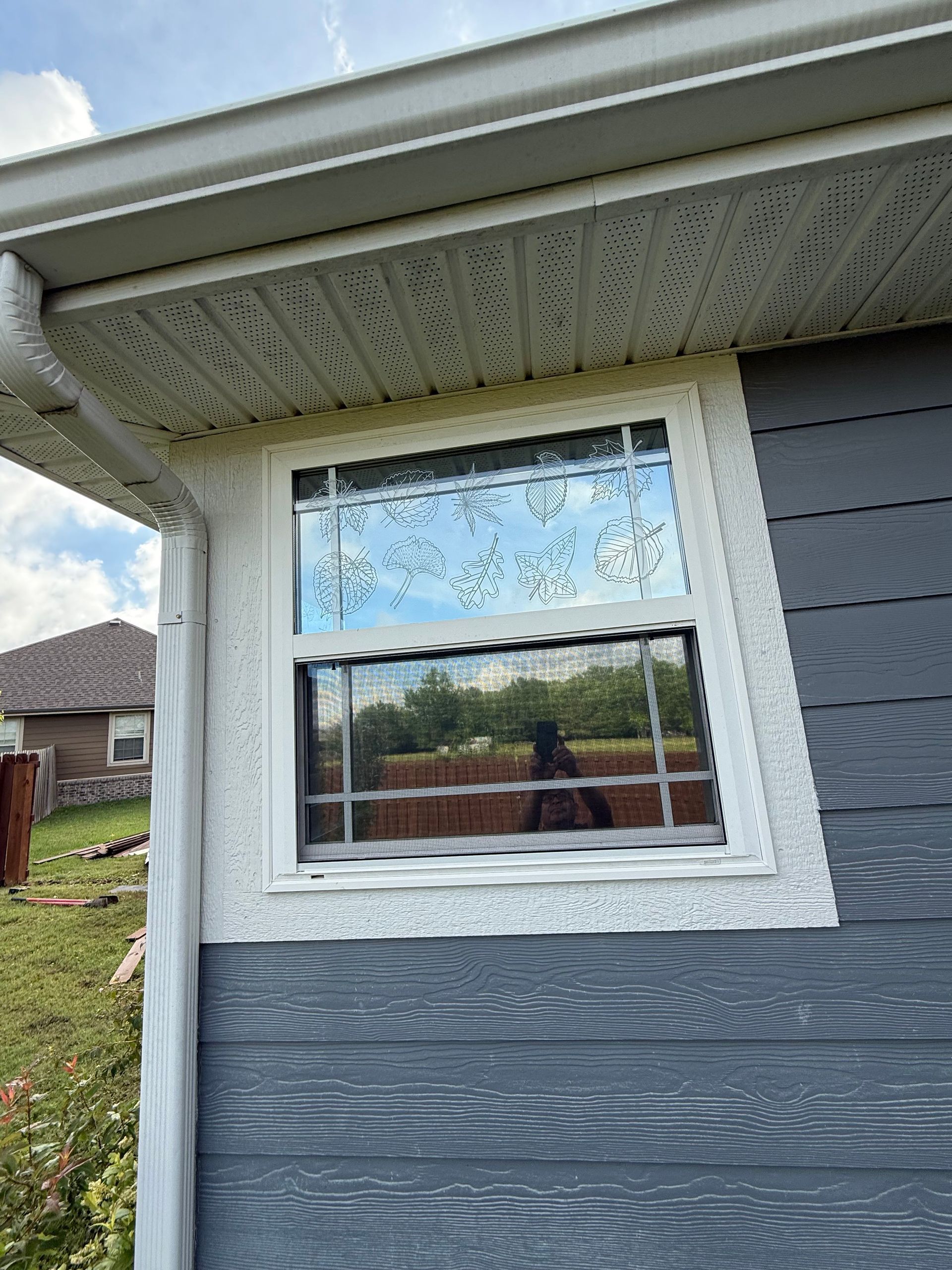 Window on a gray-sided building reflecting sky and trees. White trim and beige soffit.