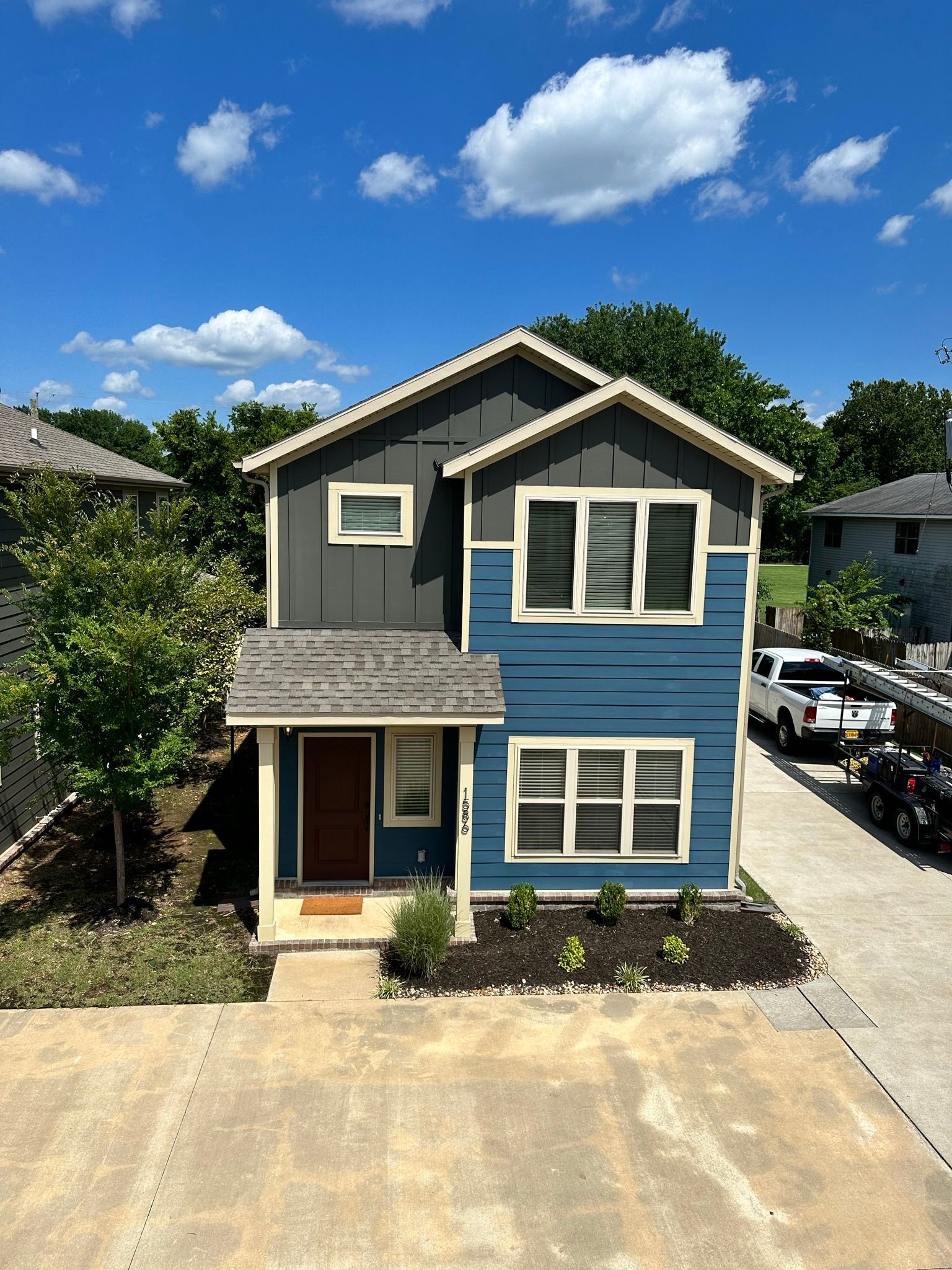 Two-story house with blue and gray siding, brown door, and small front yard with lush greenery under a blue sky.