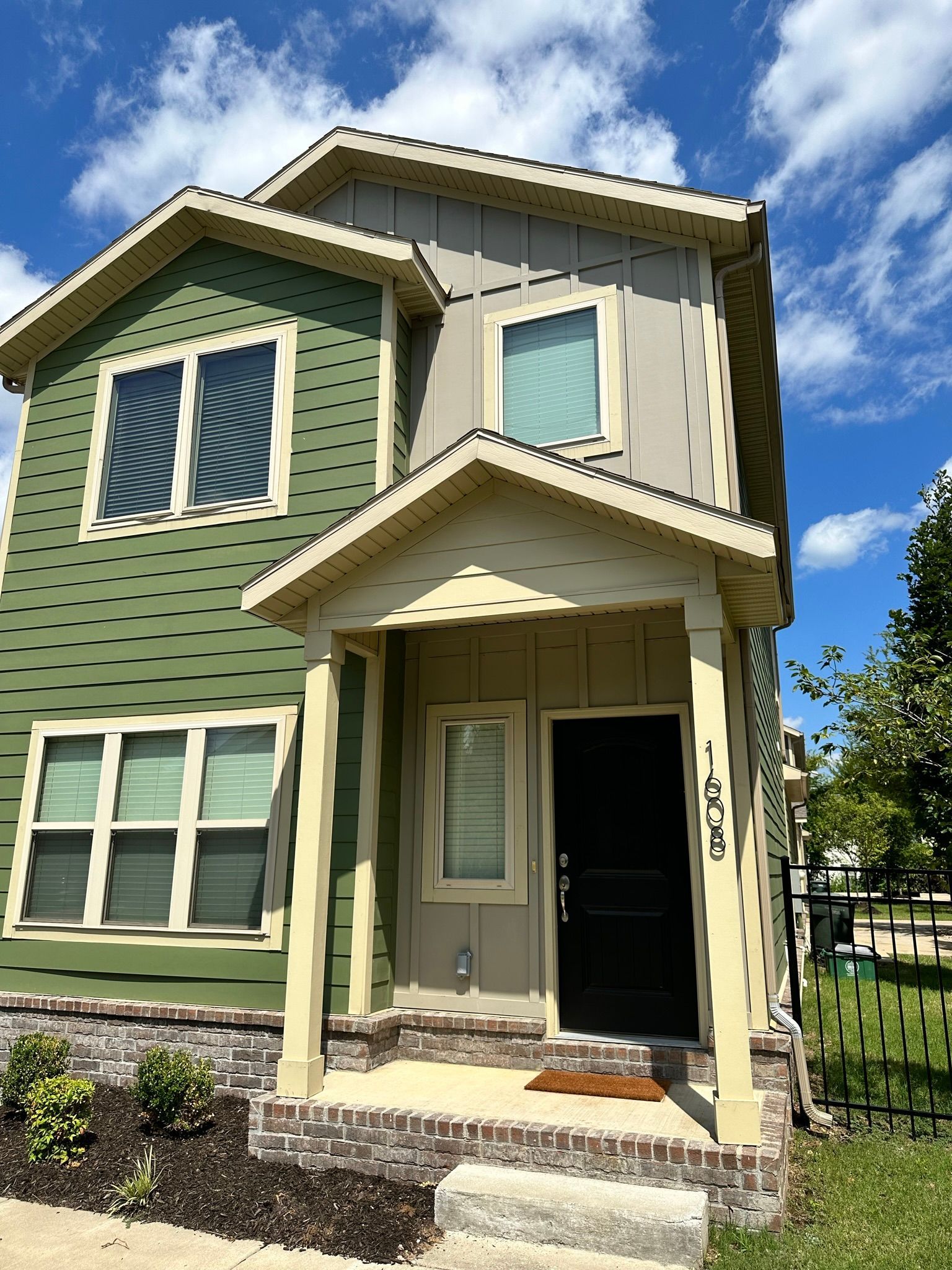 Two-story house, green and tan siding, porch, black door, small front yard, brick foundation, sunny day.