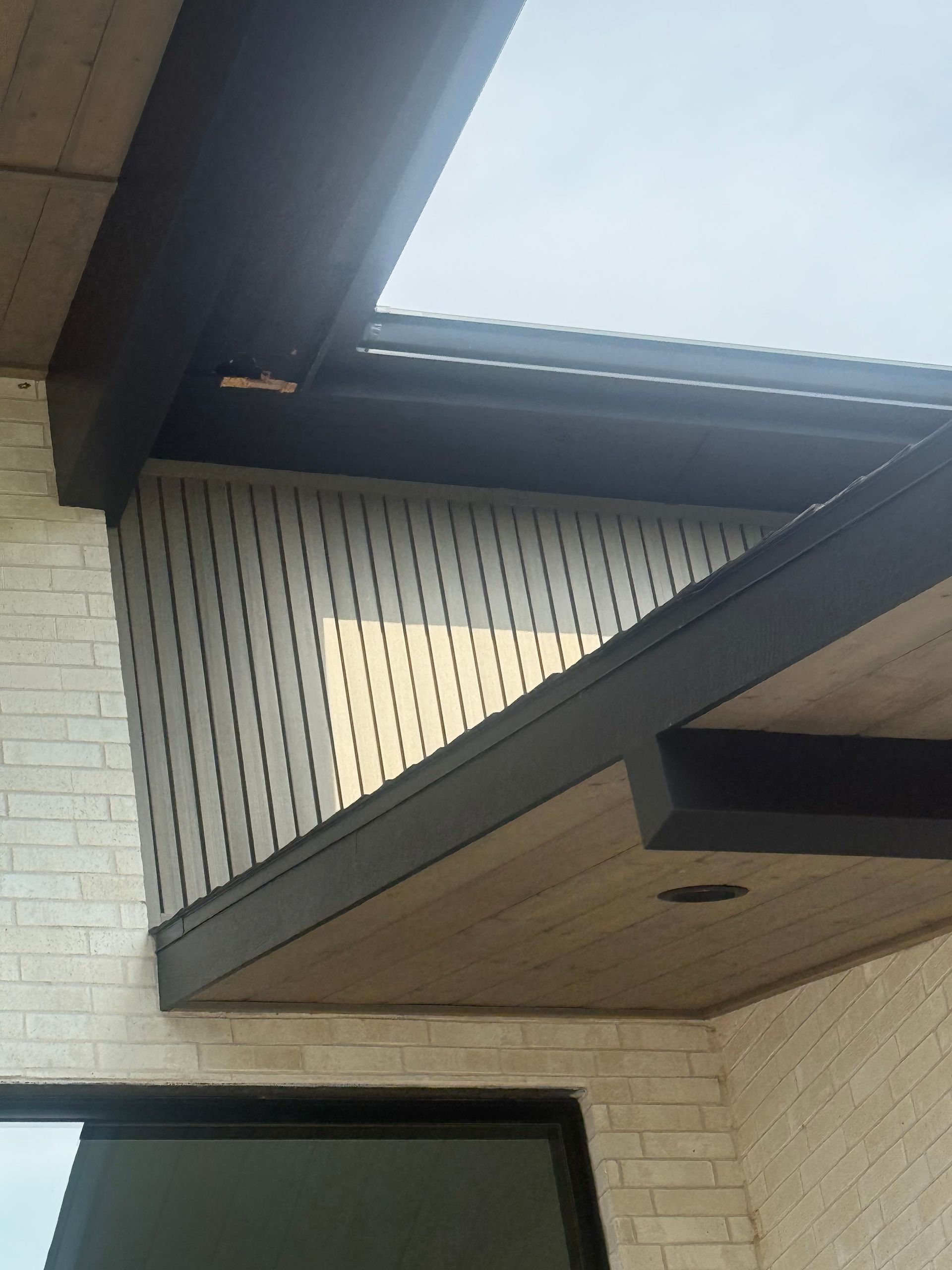 Exterior view of a building with a dark-trimmed wooden overhang, vertical siding, and light-colored brick.