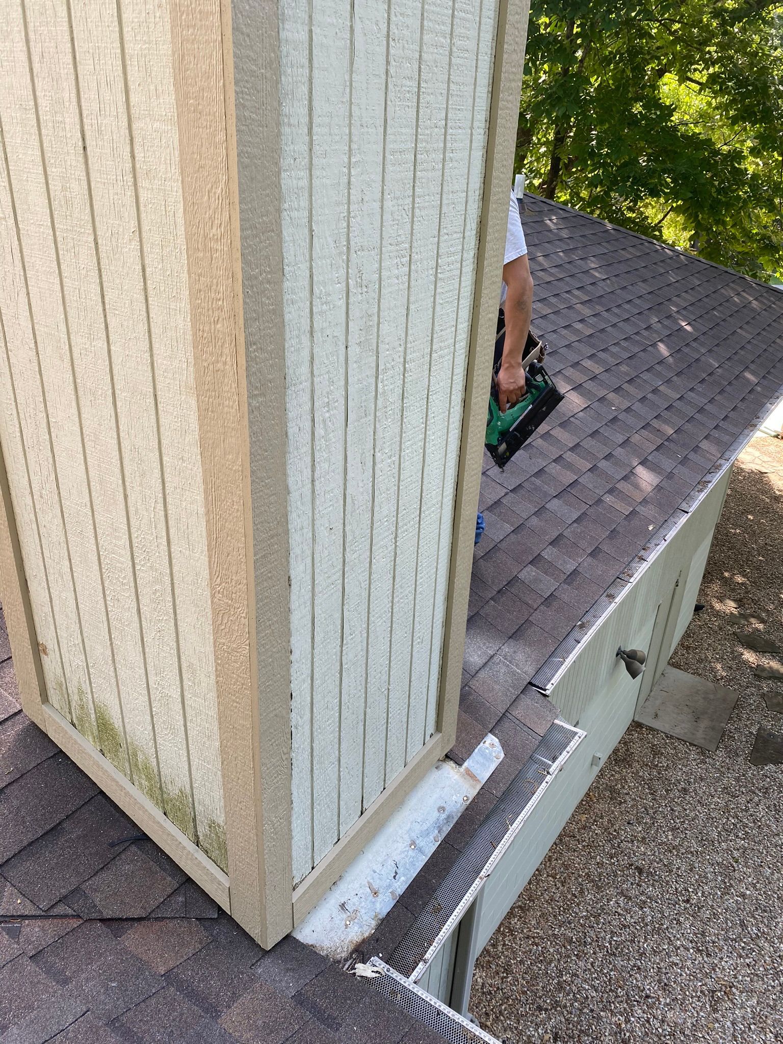 Person using a nail gun on a roof, near a light-colored structure and a dark shingled roof.