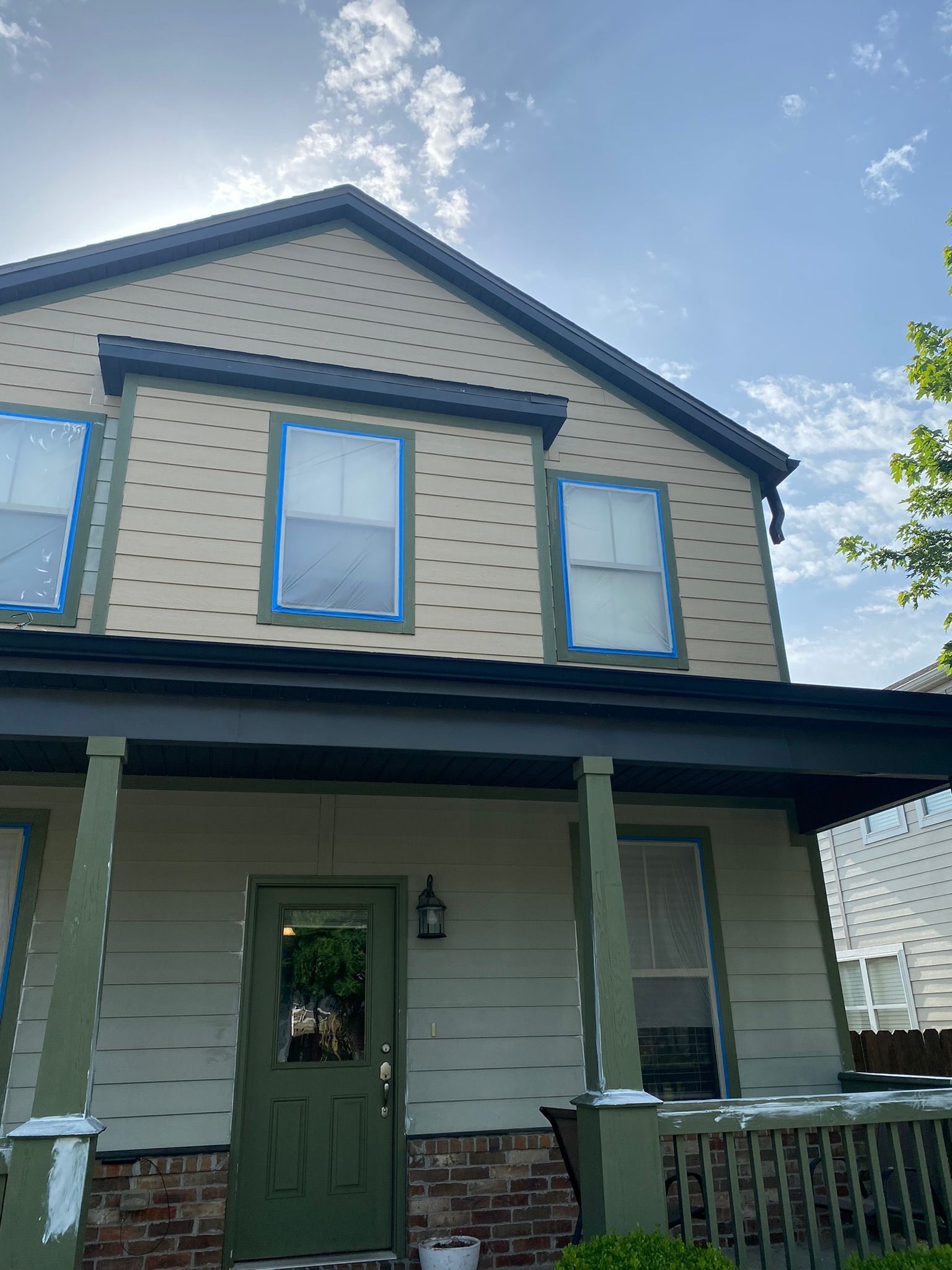 Two-story house with green and tan exterior. Windows are taped. Sunny day, blue sky.