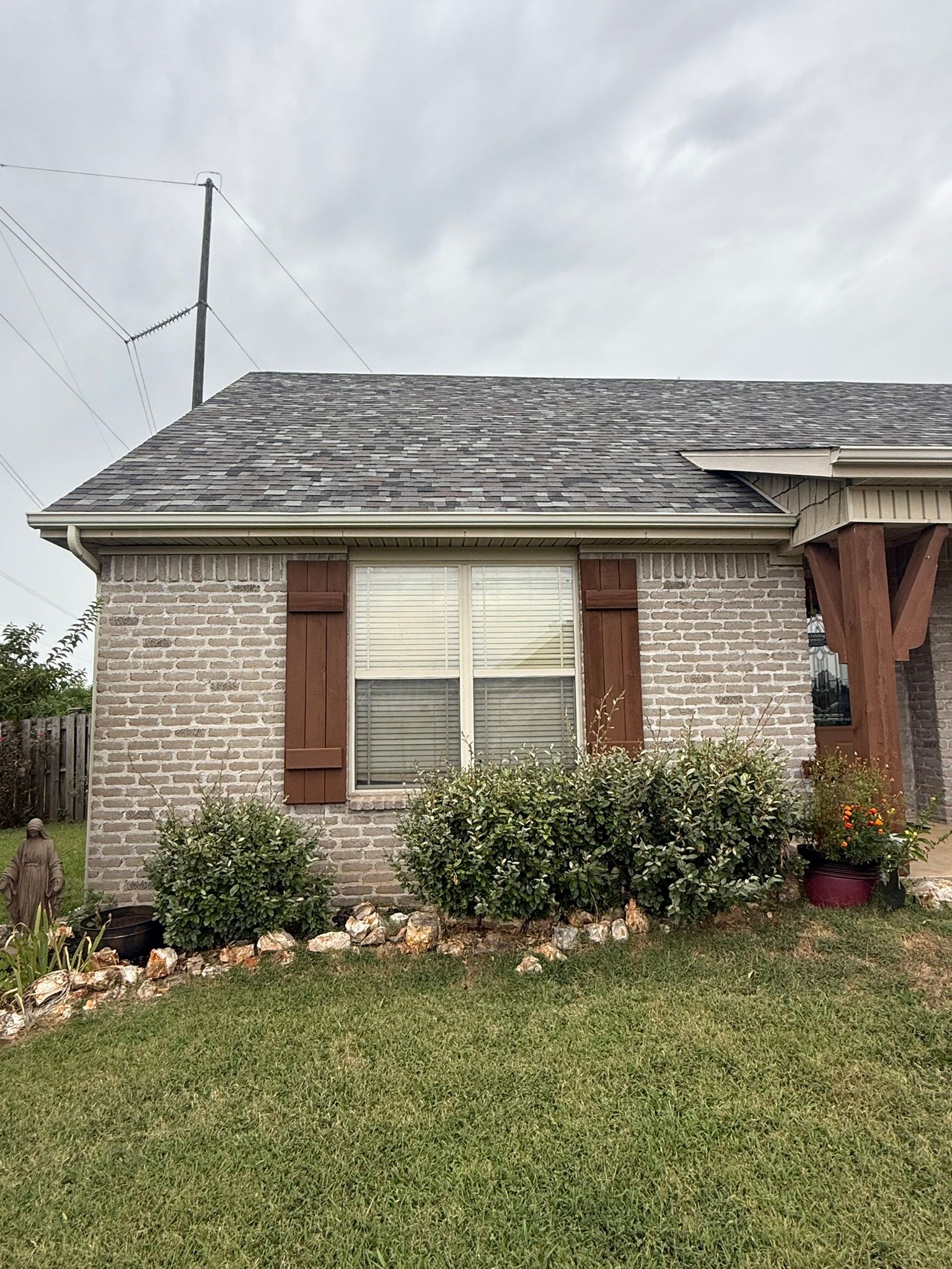 Brick house with gray roof, window with brown shutters, and shrubs in front.