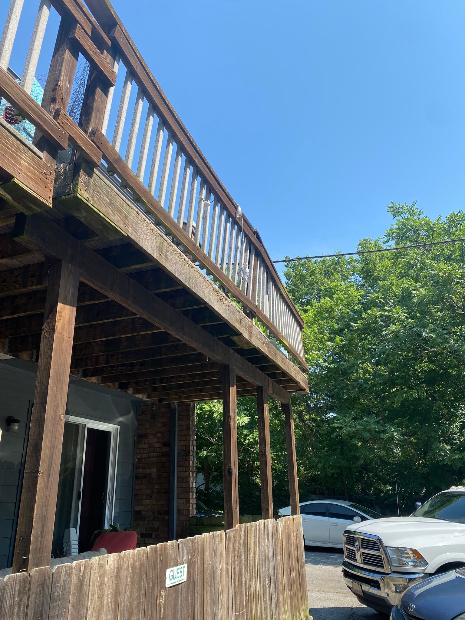 Wooden deck with railing, supported by posts, above a building with a door. Cars and green trees in the background.