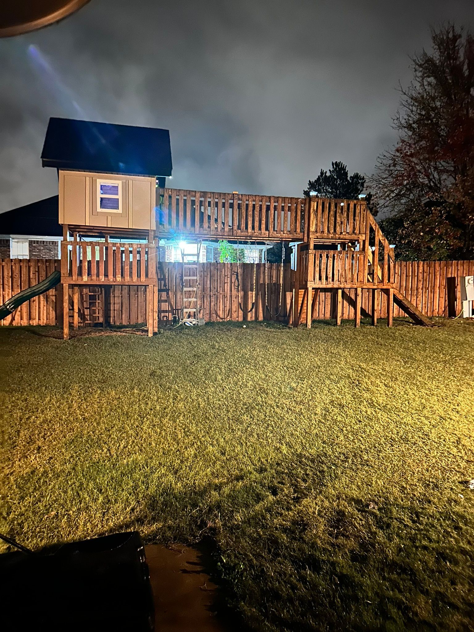 Wooden playset in a backyard at night, illuminated by soft light. Cloudy sky above.