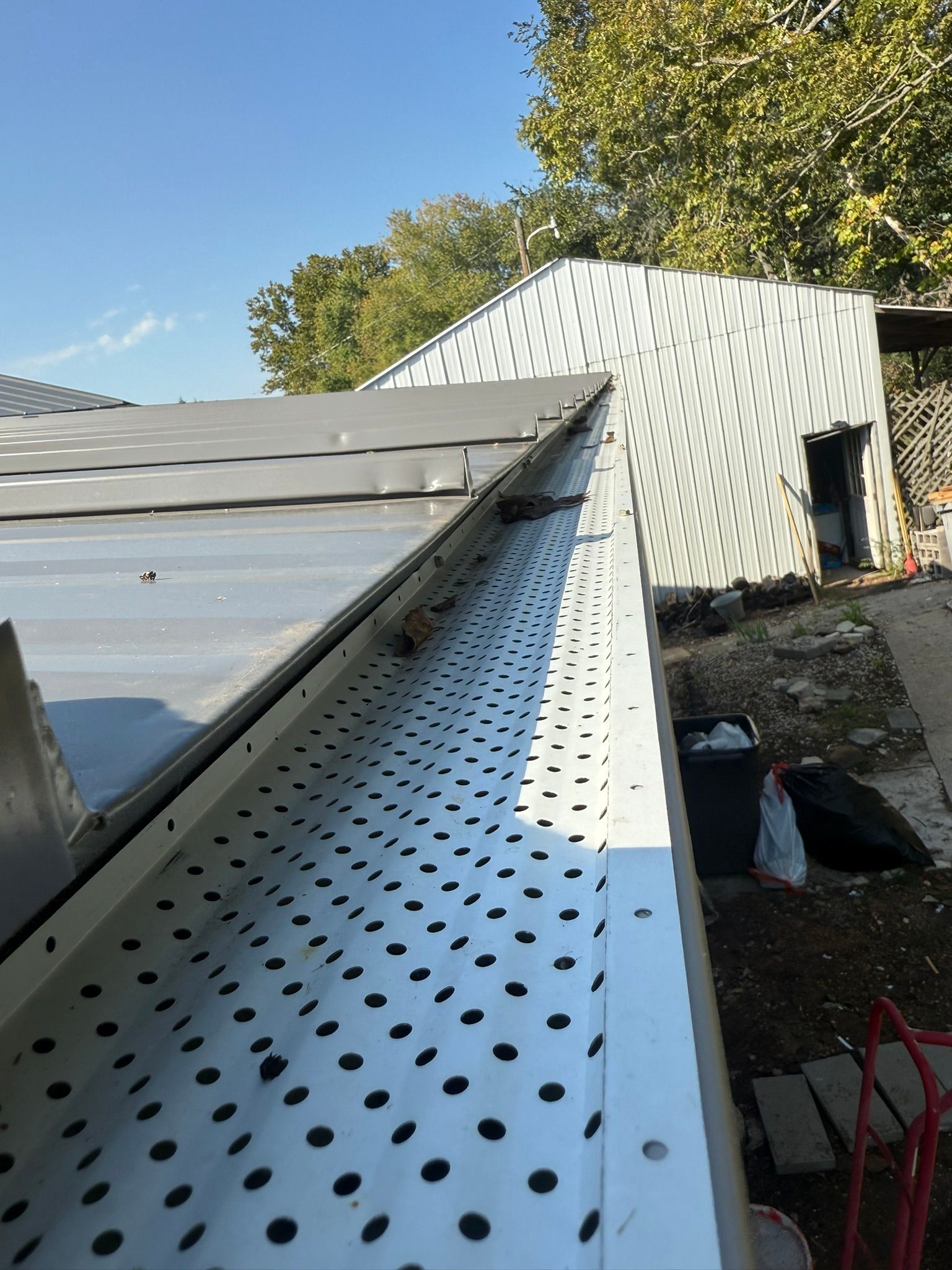 Gutter with perforated cover on a metal roof, angled view. Building exterior with trees and blue sky.