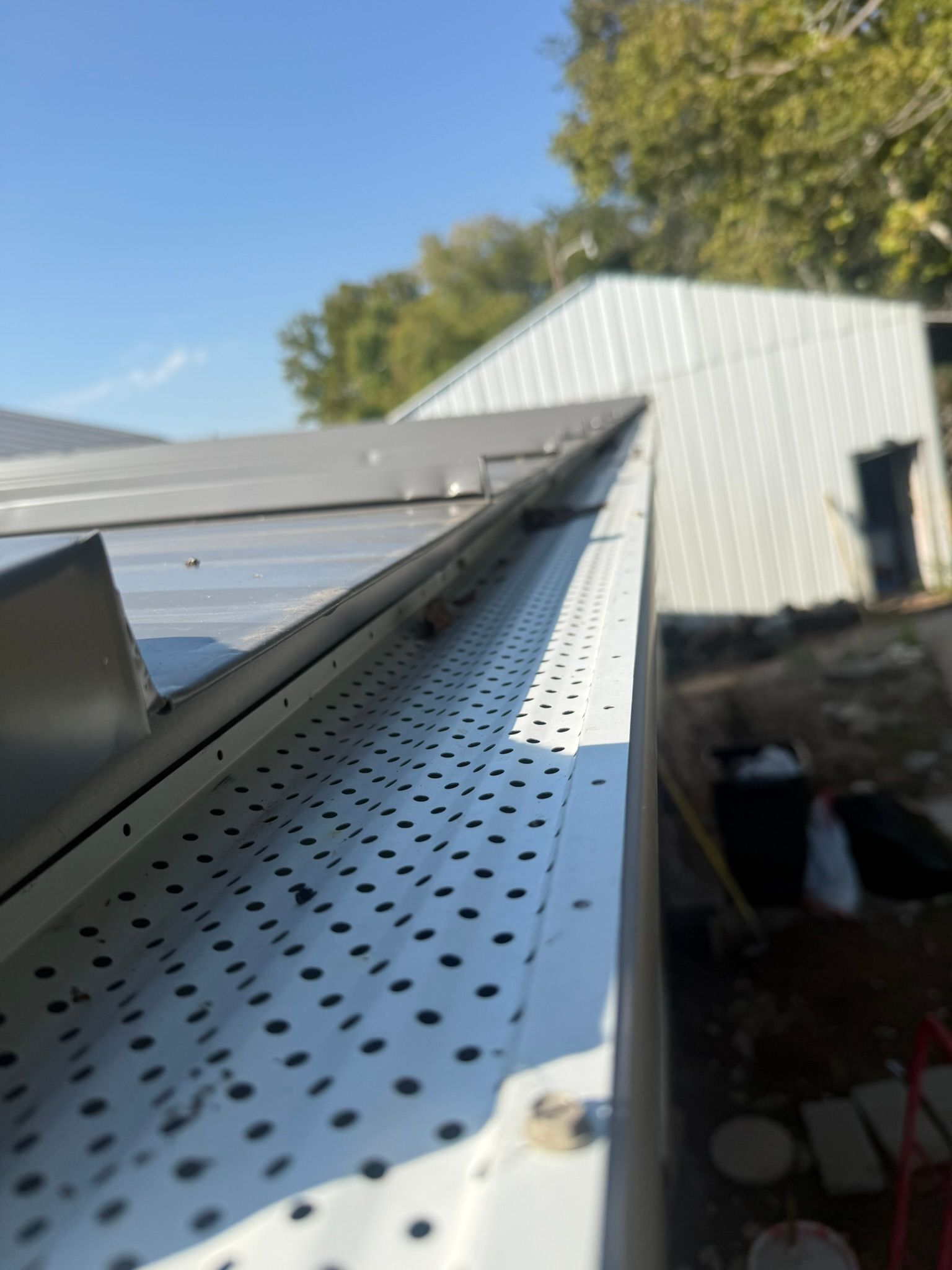 Close-up of a white gutter guard with a perforated pattern, debris visible inside; a metal building in the background.