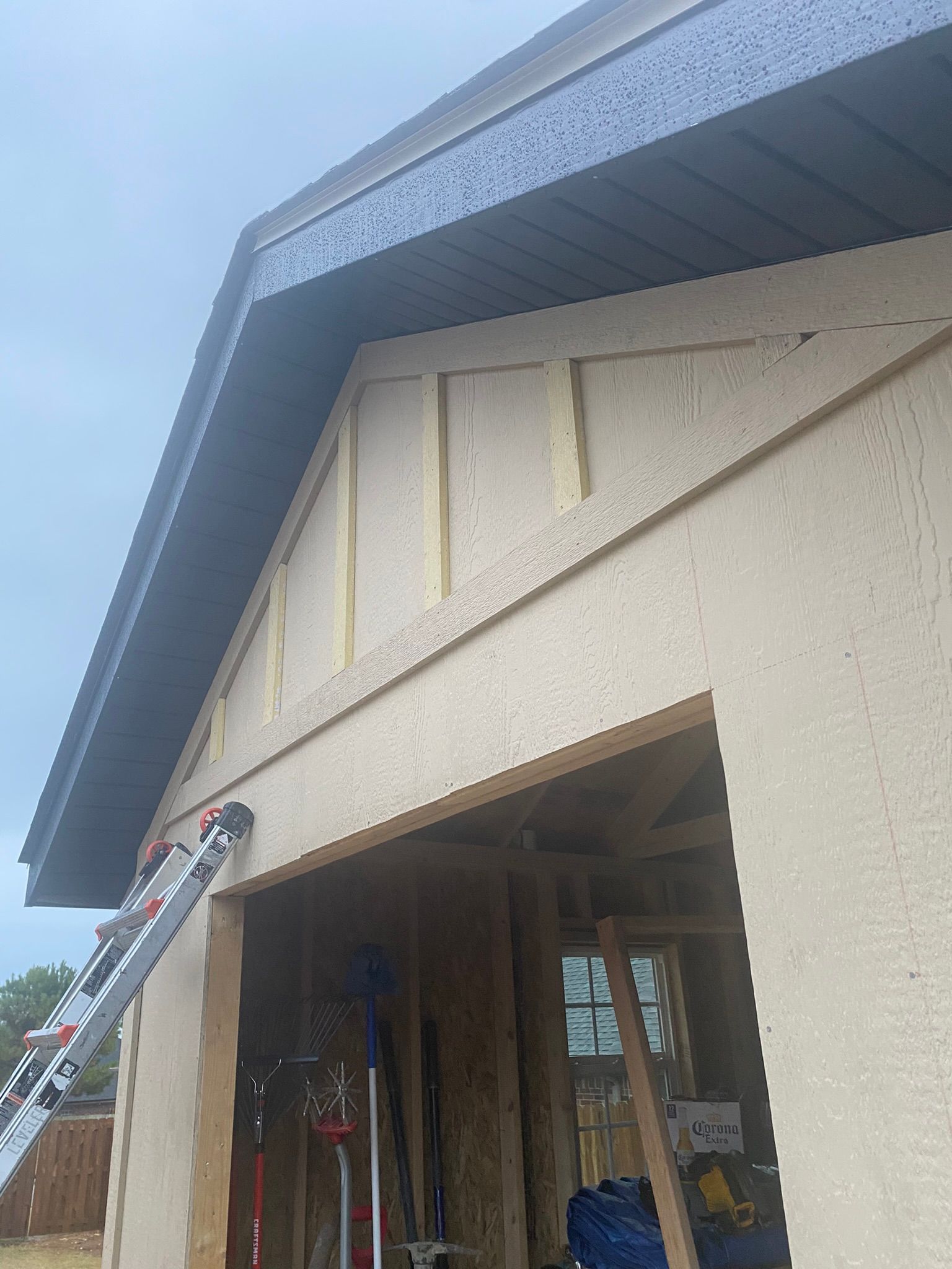 Exterior view of a building under construction, featuring a tan stucco facade, a dark roof, and a ladder.