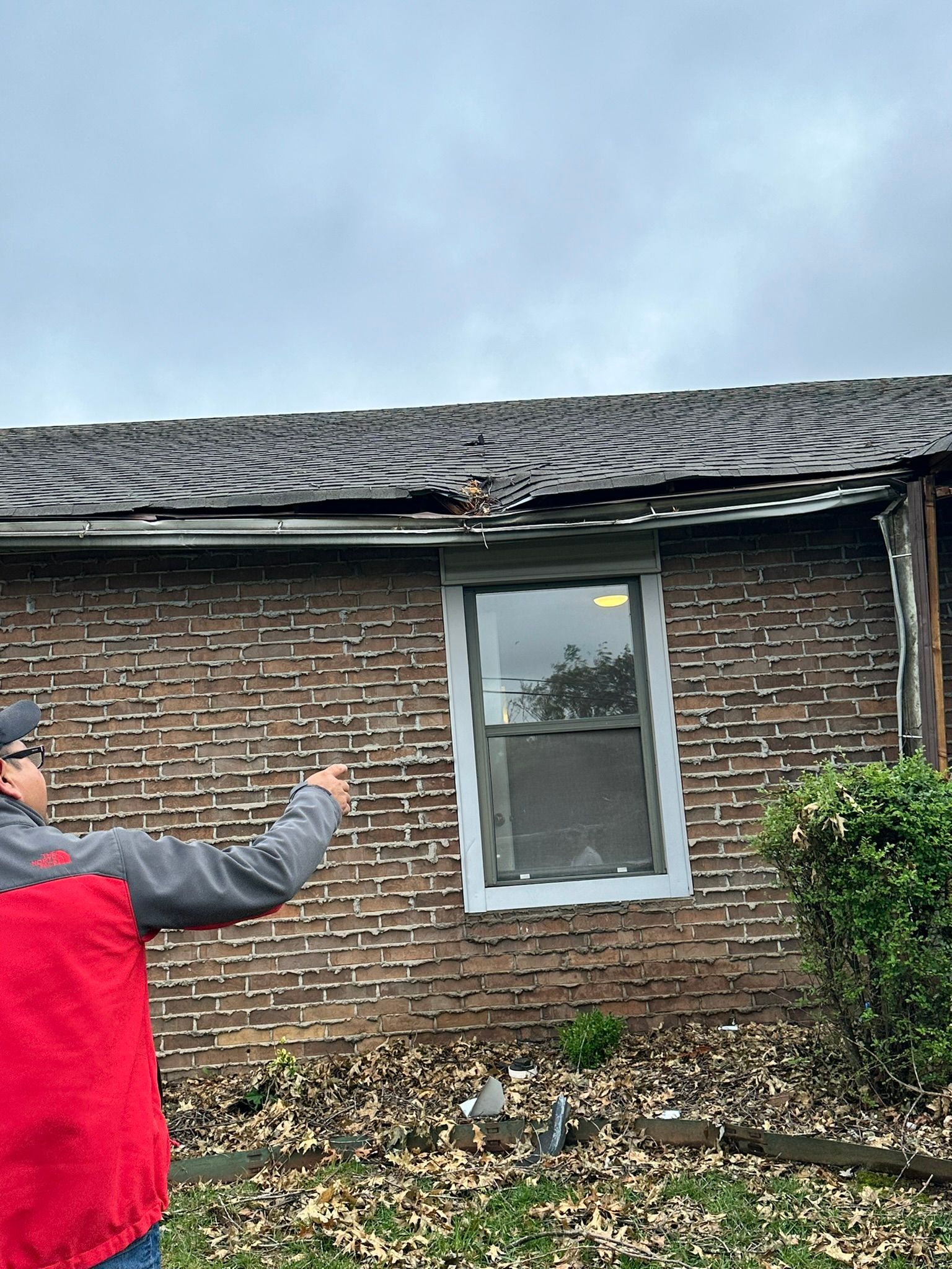 Person pointing at damaged roof of a brick building near a window. Overcast sky.