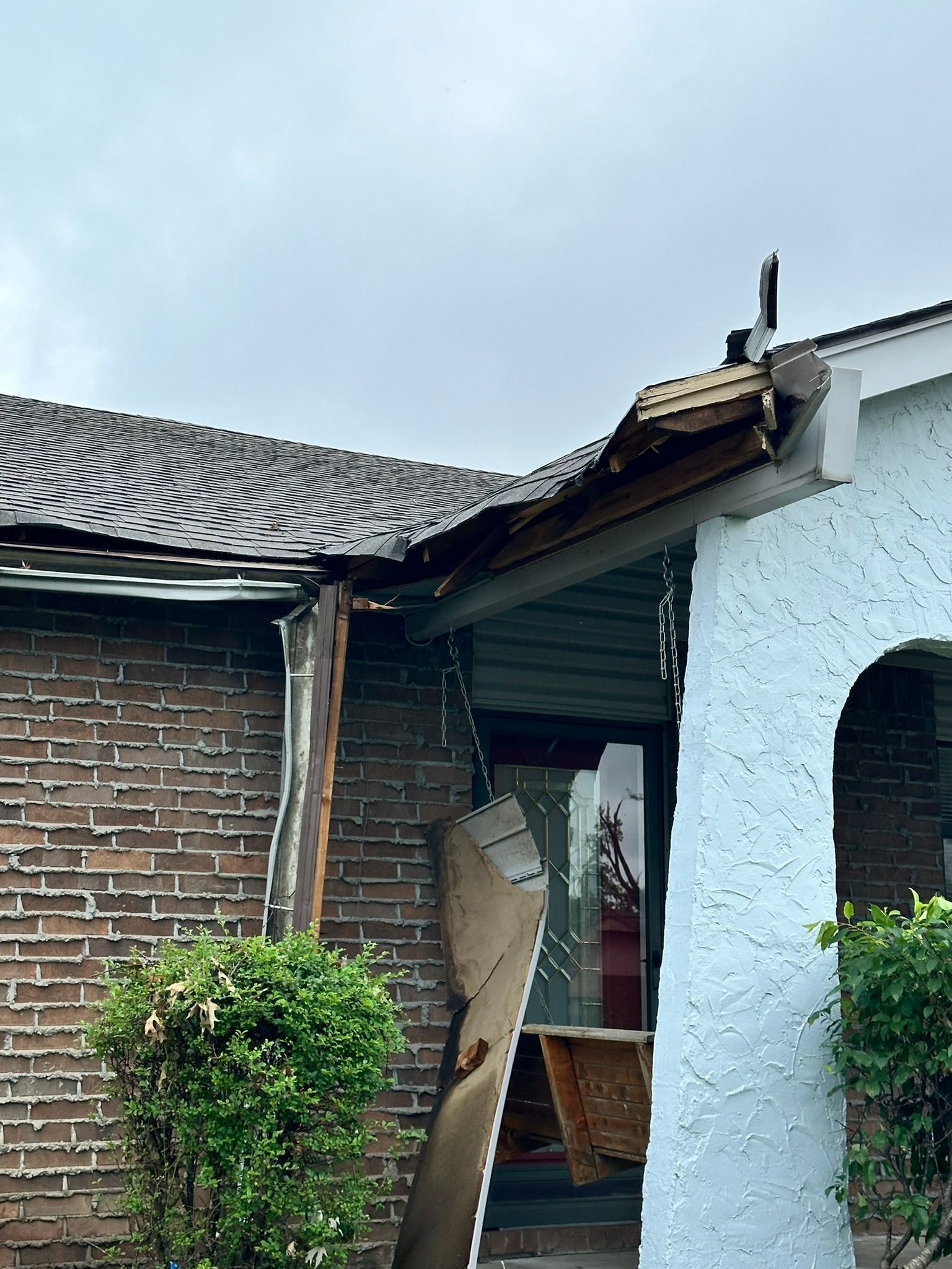 Damaged home exterior showing missing roof sections, exposed wood, and structural damage near a door.