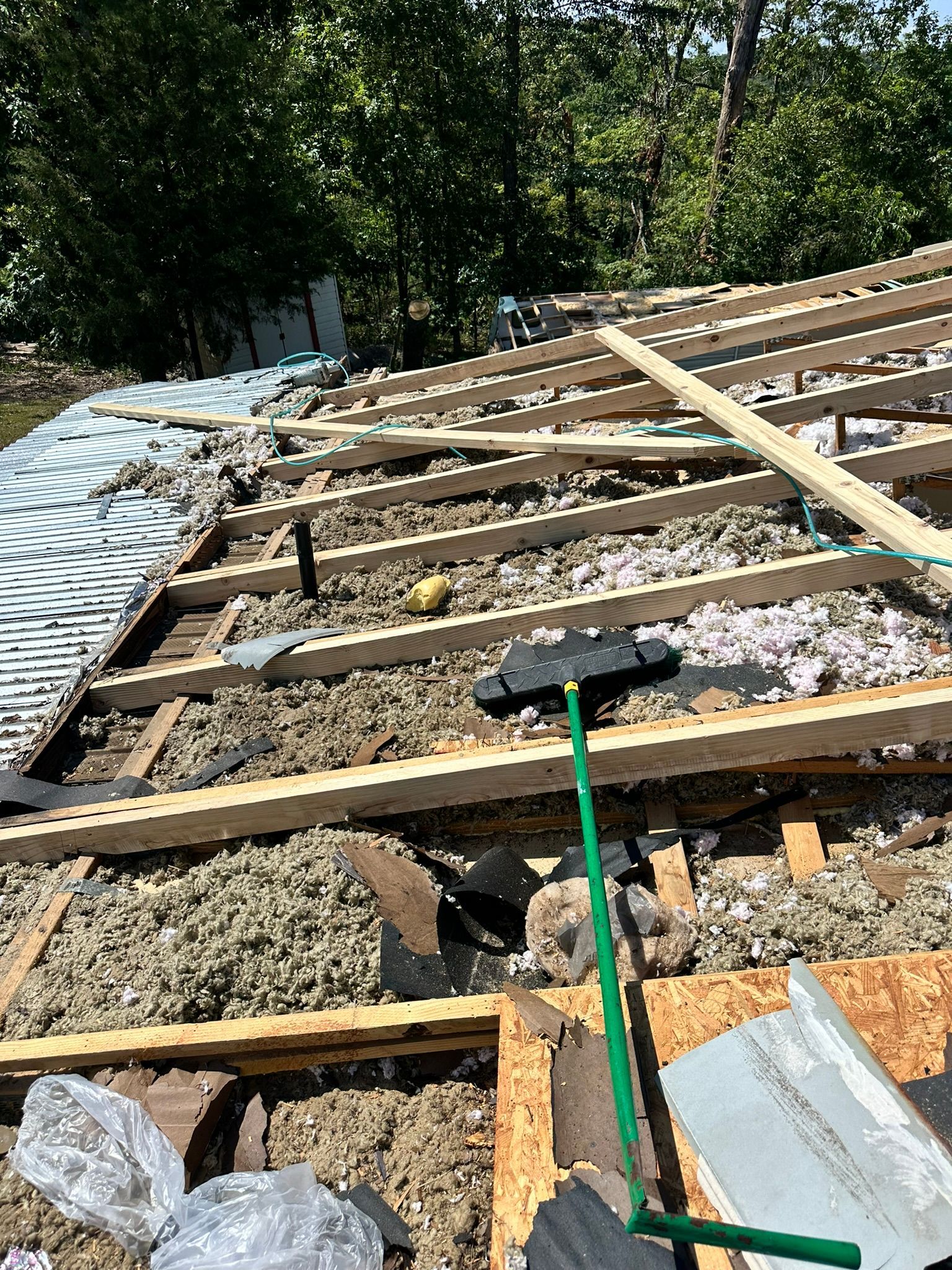 Roof partially dismantled, insulation exposed. A green-handled brush rests on the roof. Sunlight, trees in the background.