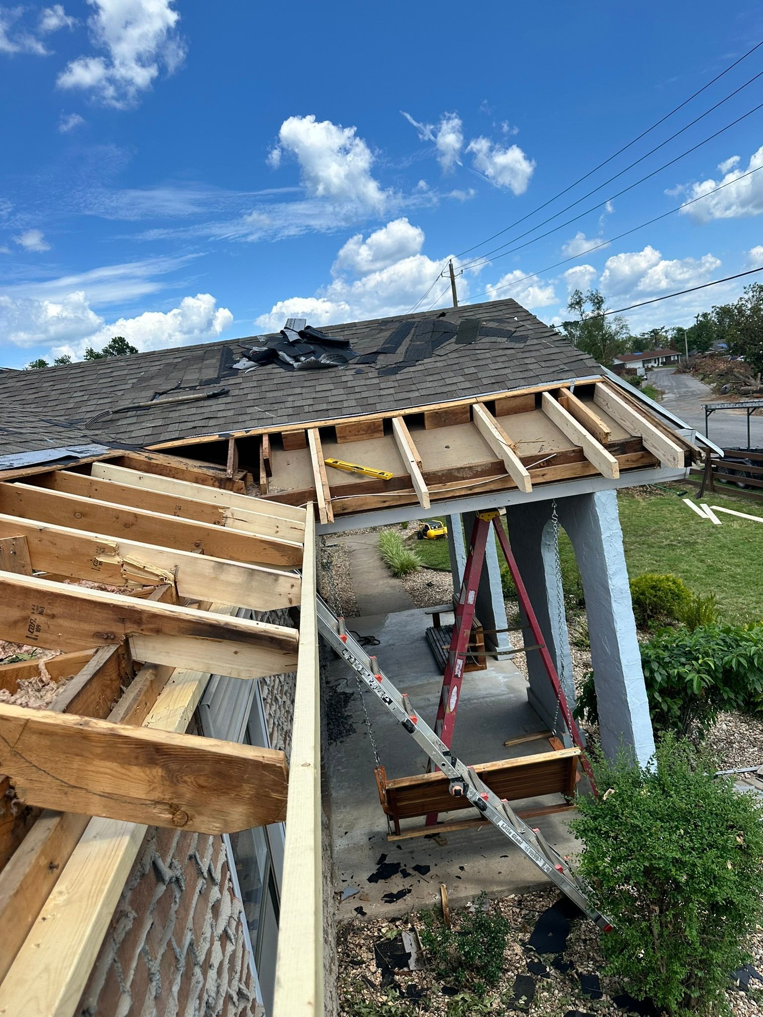House roof damaged, showing exposed rafters and missing shingles. Blue sky and sunny day.