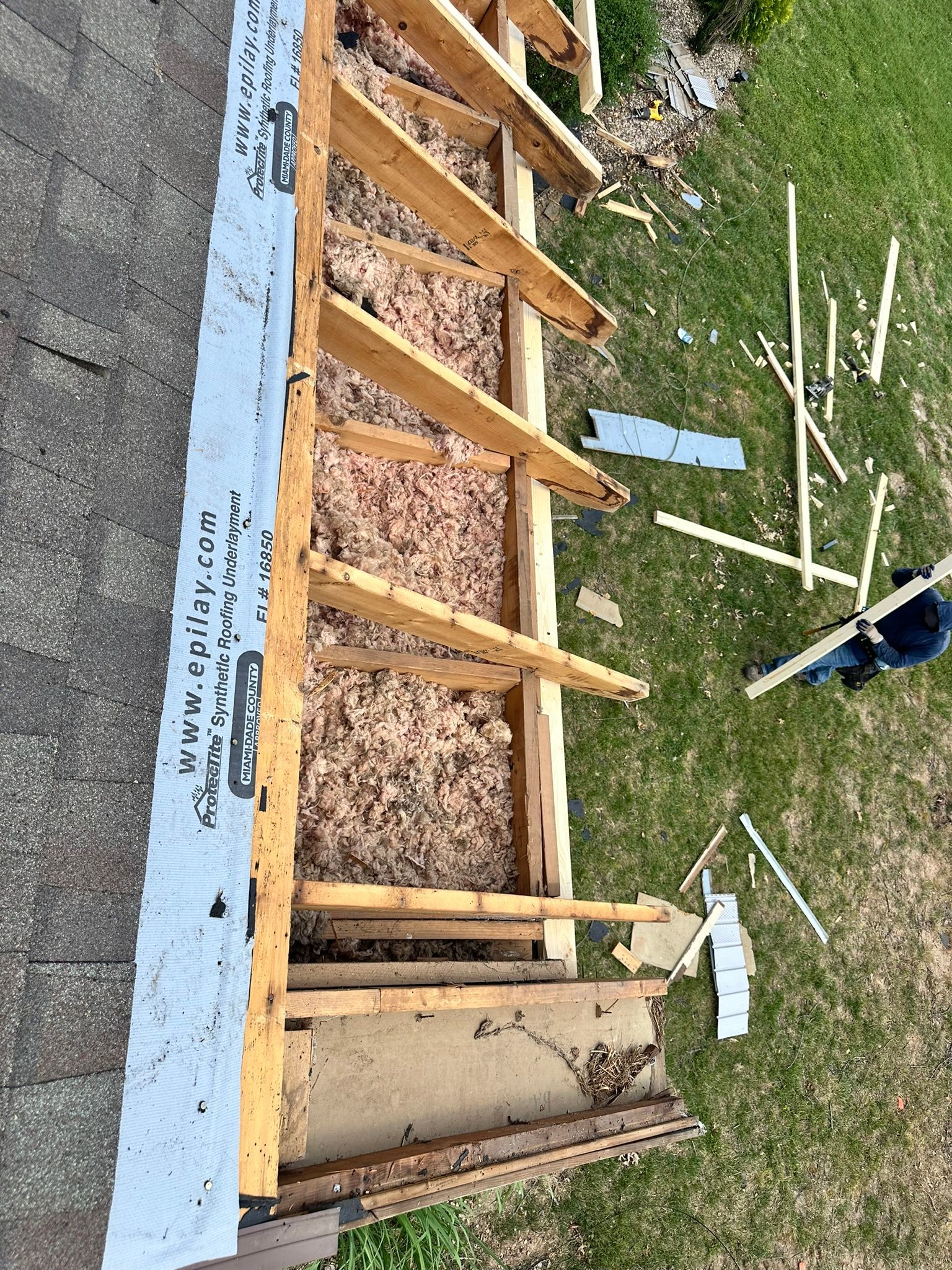 Rooftop with exposed wooden beams, insulation, and building materials; person in blue on the right side of the lawn.