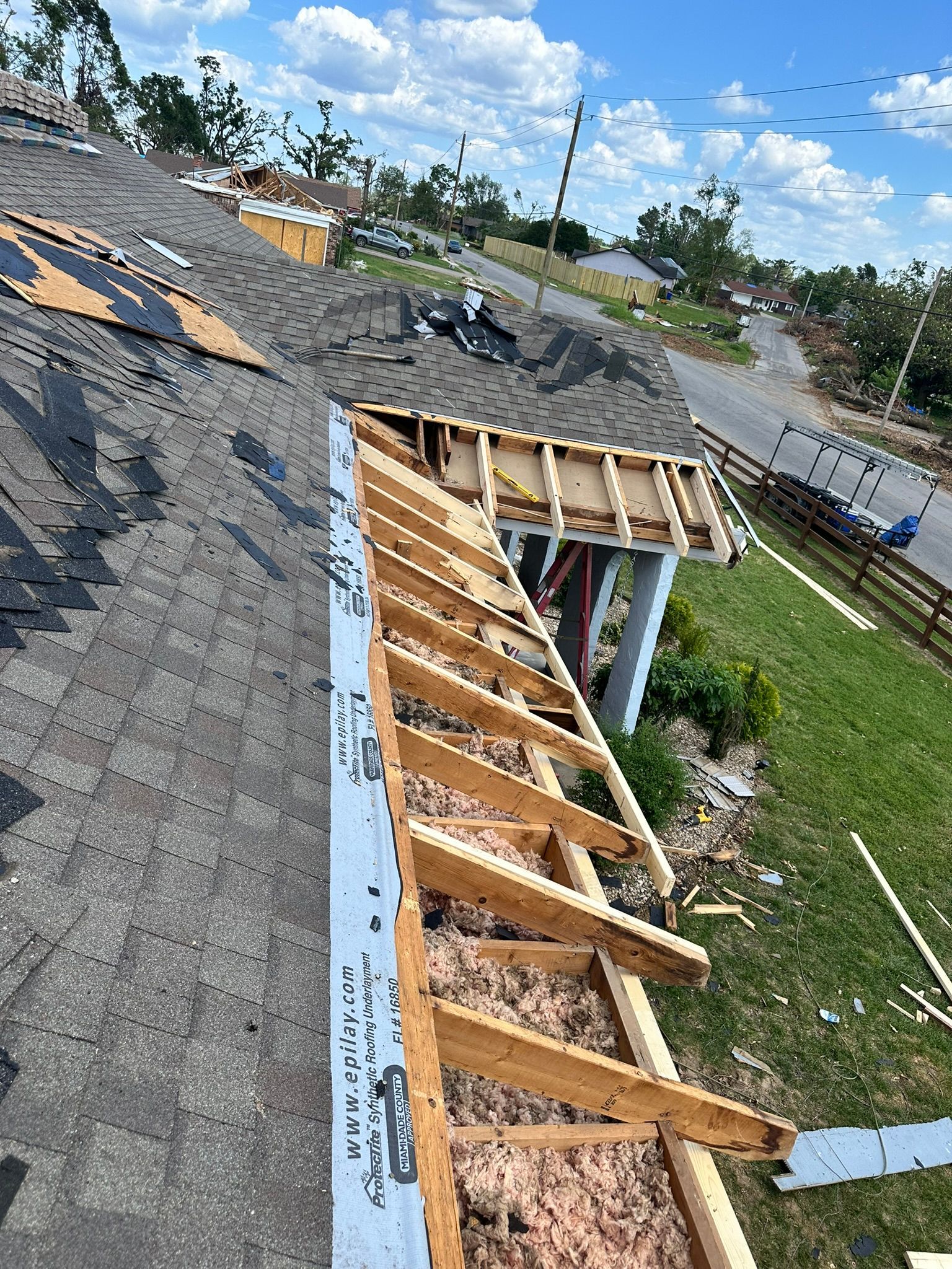 Damaged house roof with exposed wood framing, insulation, and missing shingles. Debris scattered nearby.