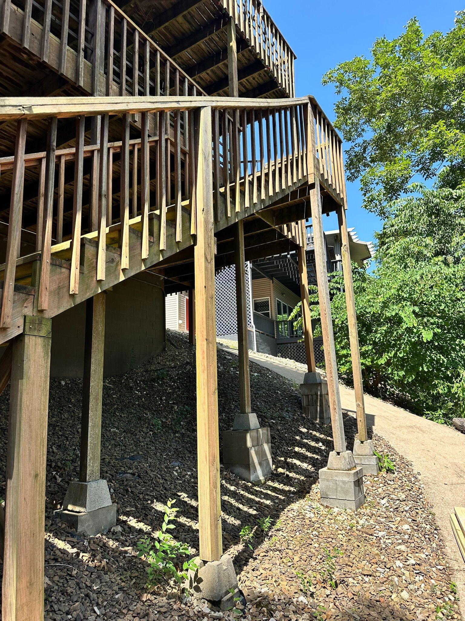 Wooden deck with stairs, supported by posts on concrete bases, overlooking a gravel-covered hillside.