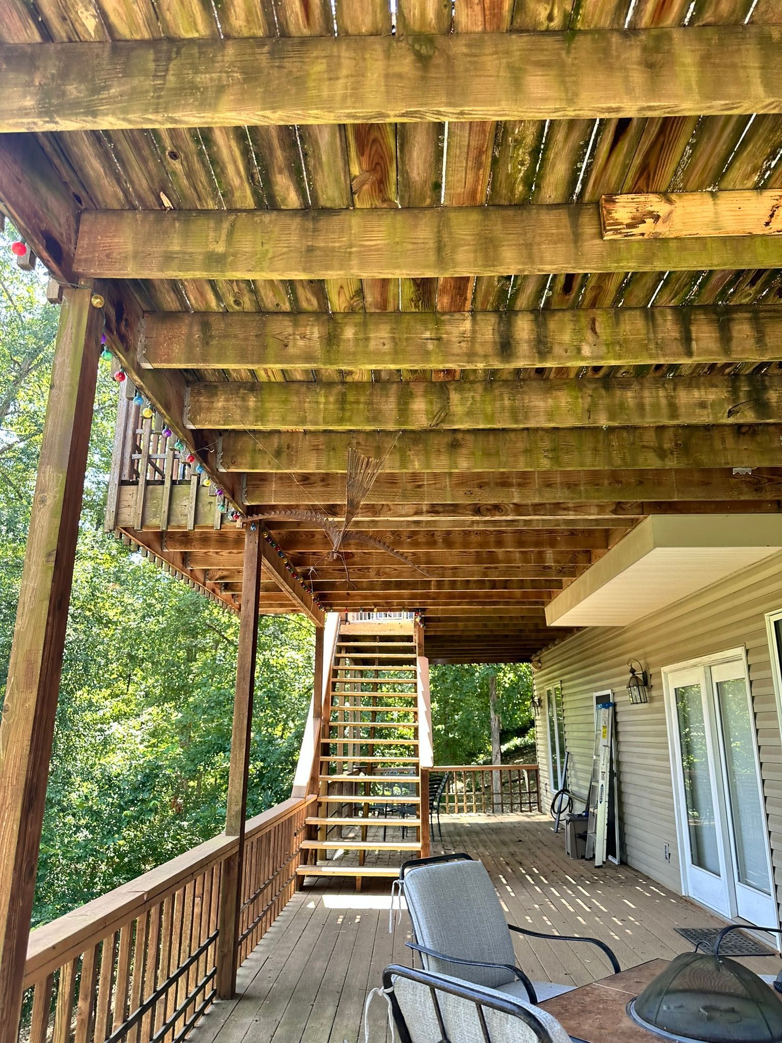 Wooden deck with stairs, under deck view. Green and brown tones. Sunlight and trees in background.
