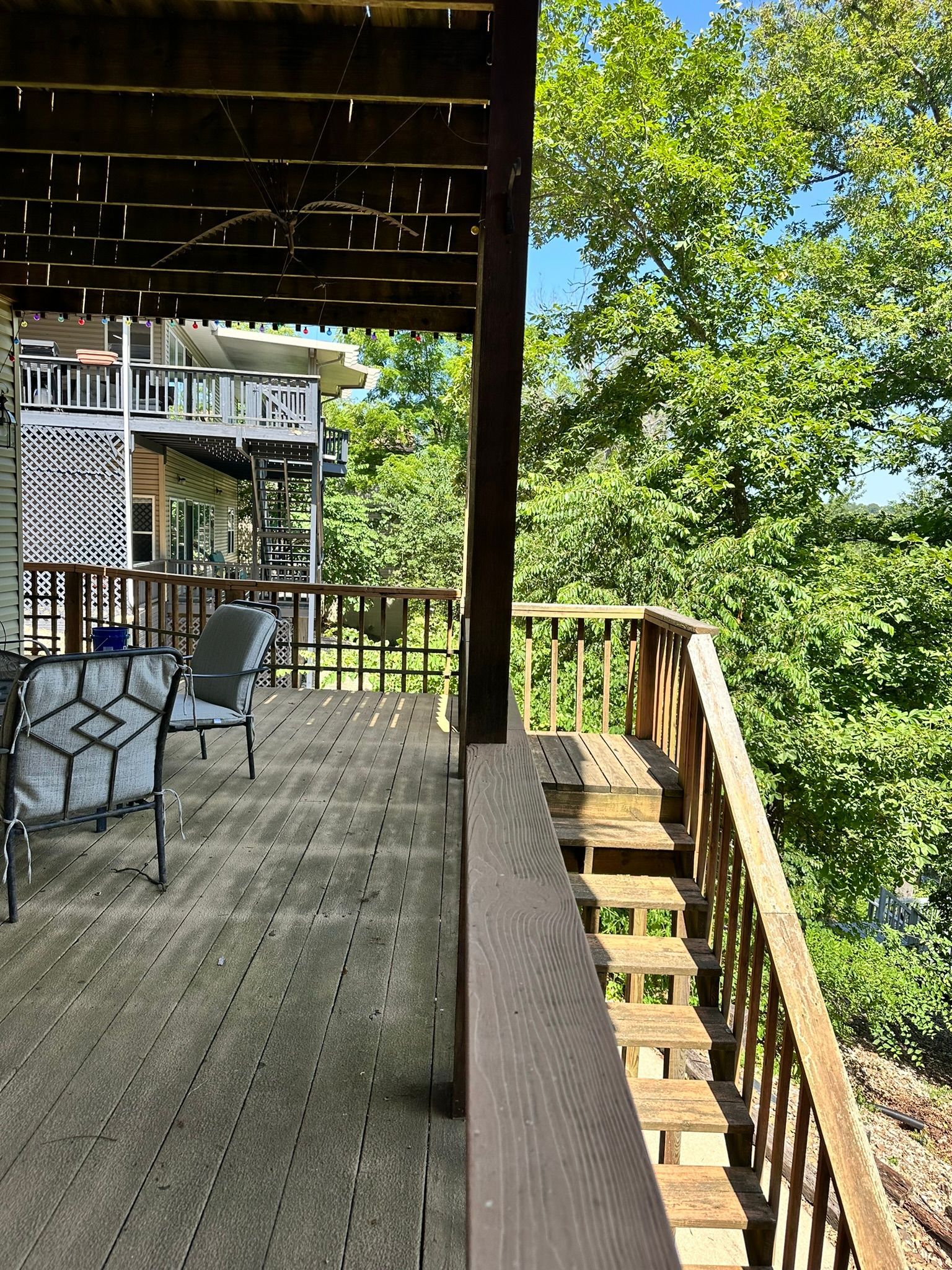 Wooden deck with stairs, surrounded by railing. Trees and another deck visible in the background.