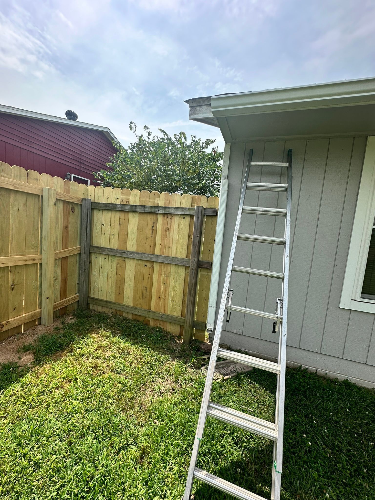 A wooden fence with a ladder leaning against a light-colored house. Green grass and a cloudy sky.