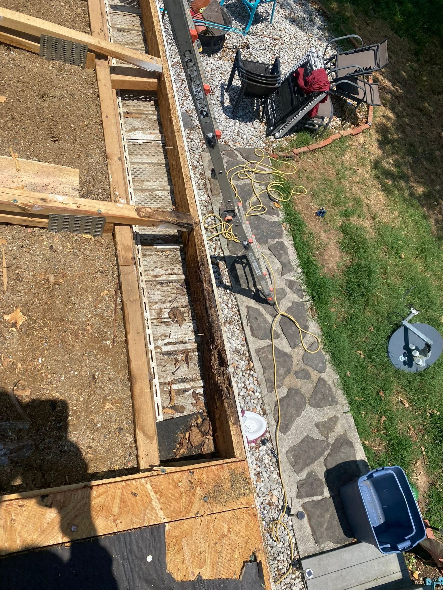 Roof with exposed wood framing, debris, and tools on the edge; patio with stone walkway and yard.