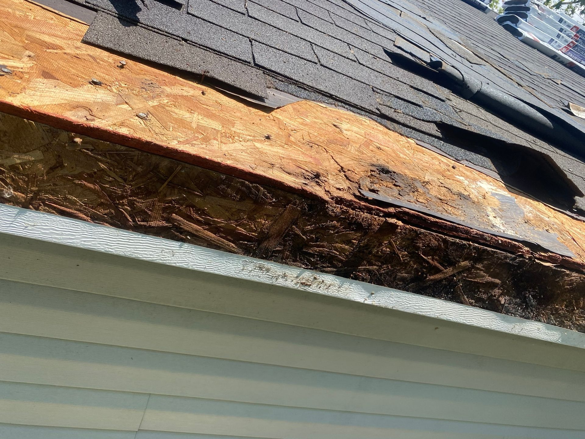 Damaged roof edge with missing shingles exposing rotted wood above a gutter.