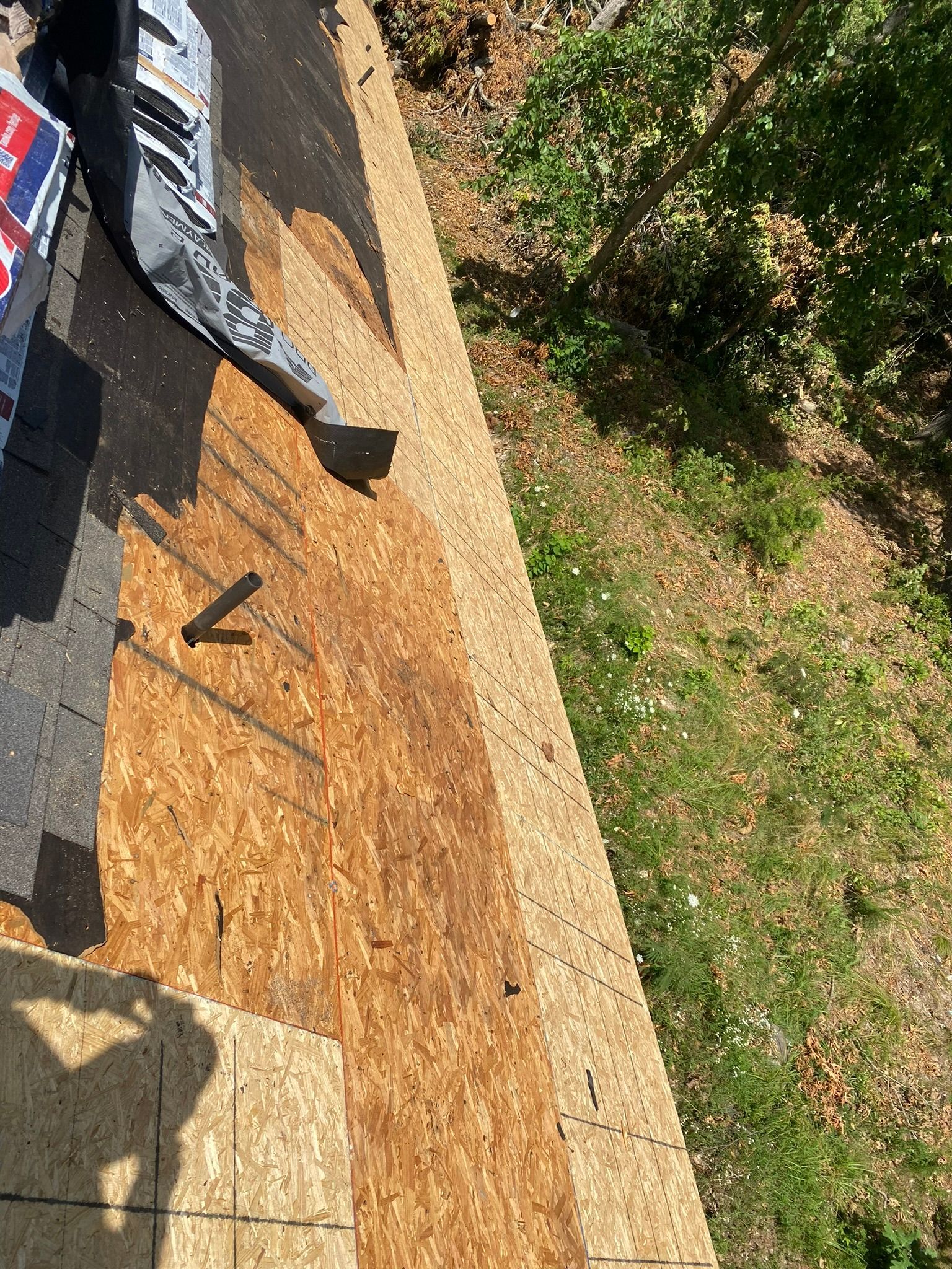 Close-up of a partially shingled roof with exposed wood sheathing. A person's legs are visible.