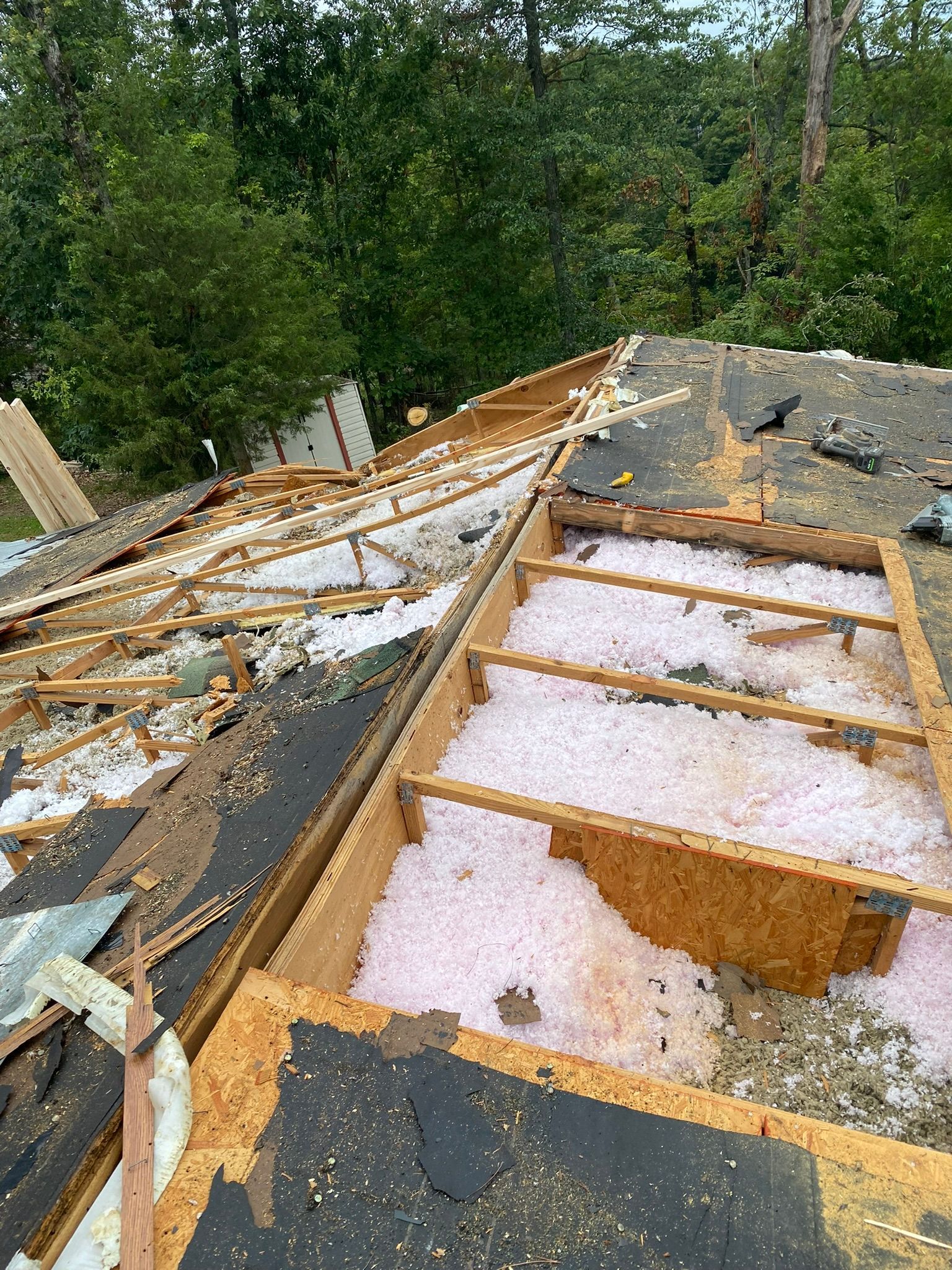 Roof partially torn off, revealing wooden framework and insulation, with trees in the background.