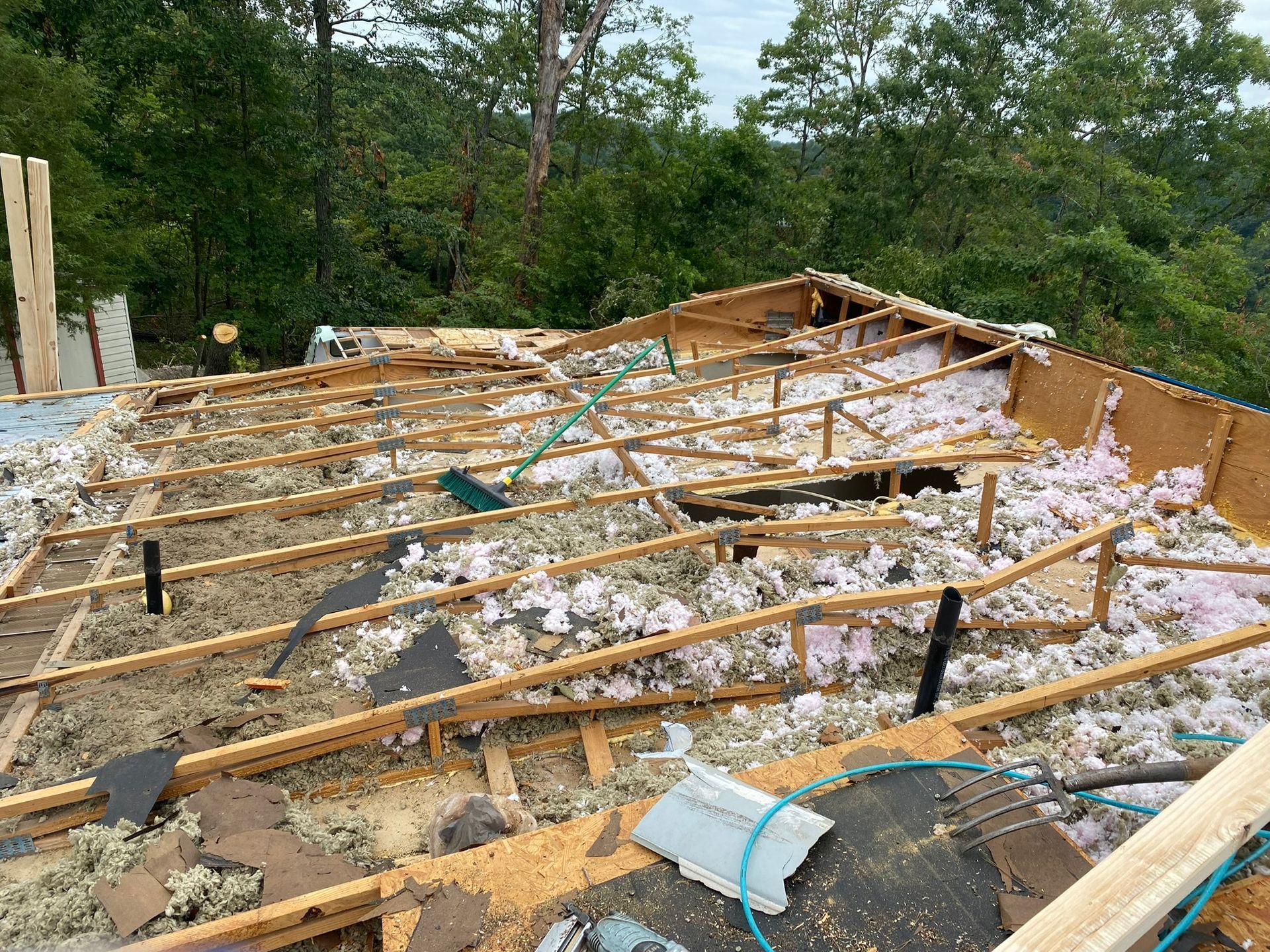 Roof under construction with exposed beams, insulation, and debris. Trees in the background.