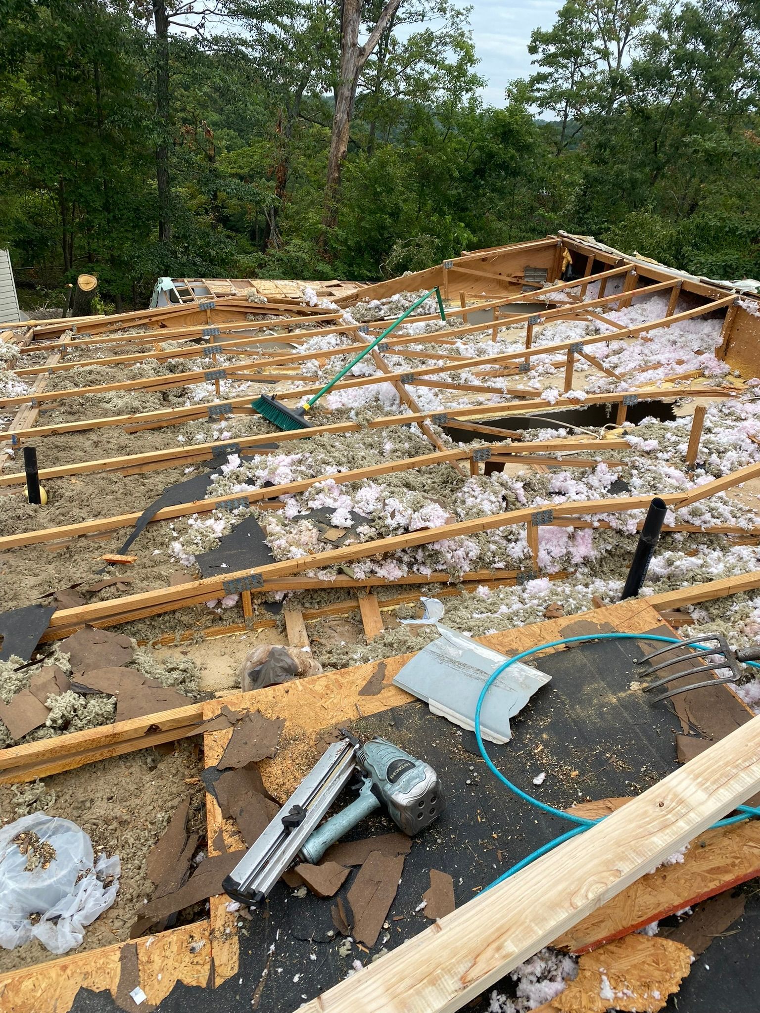 Rooftop in disrepair, wooden beams exposed, insulation visible, tools and debris scattered, background trees.