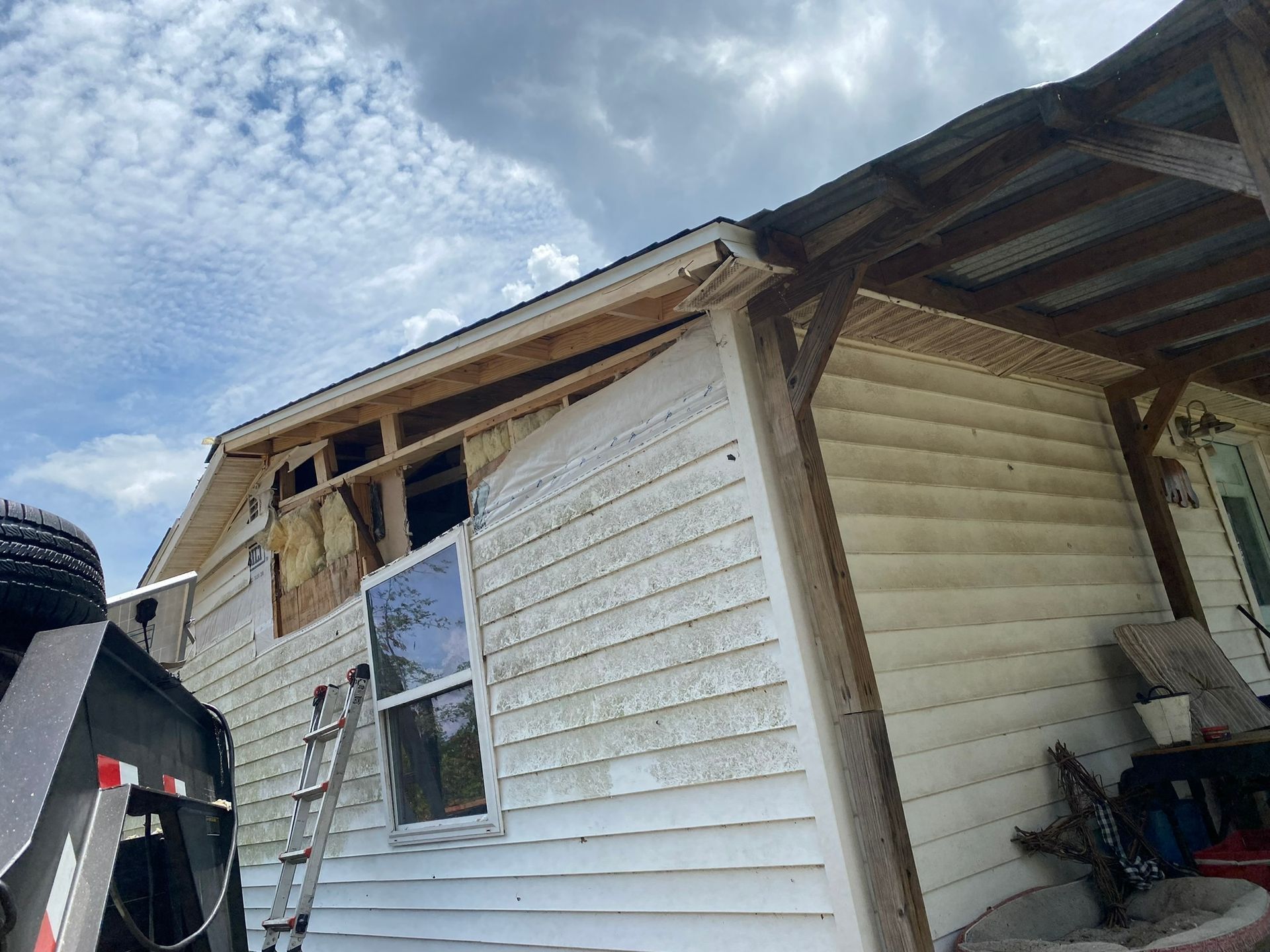 House under construction; siding partially removed, open window, ladder propped, trailer nearby.