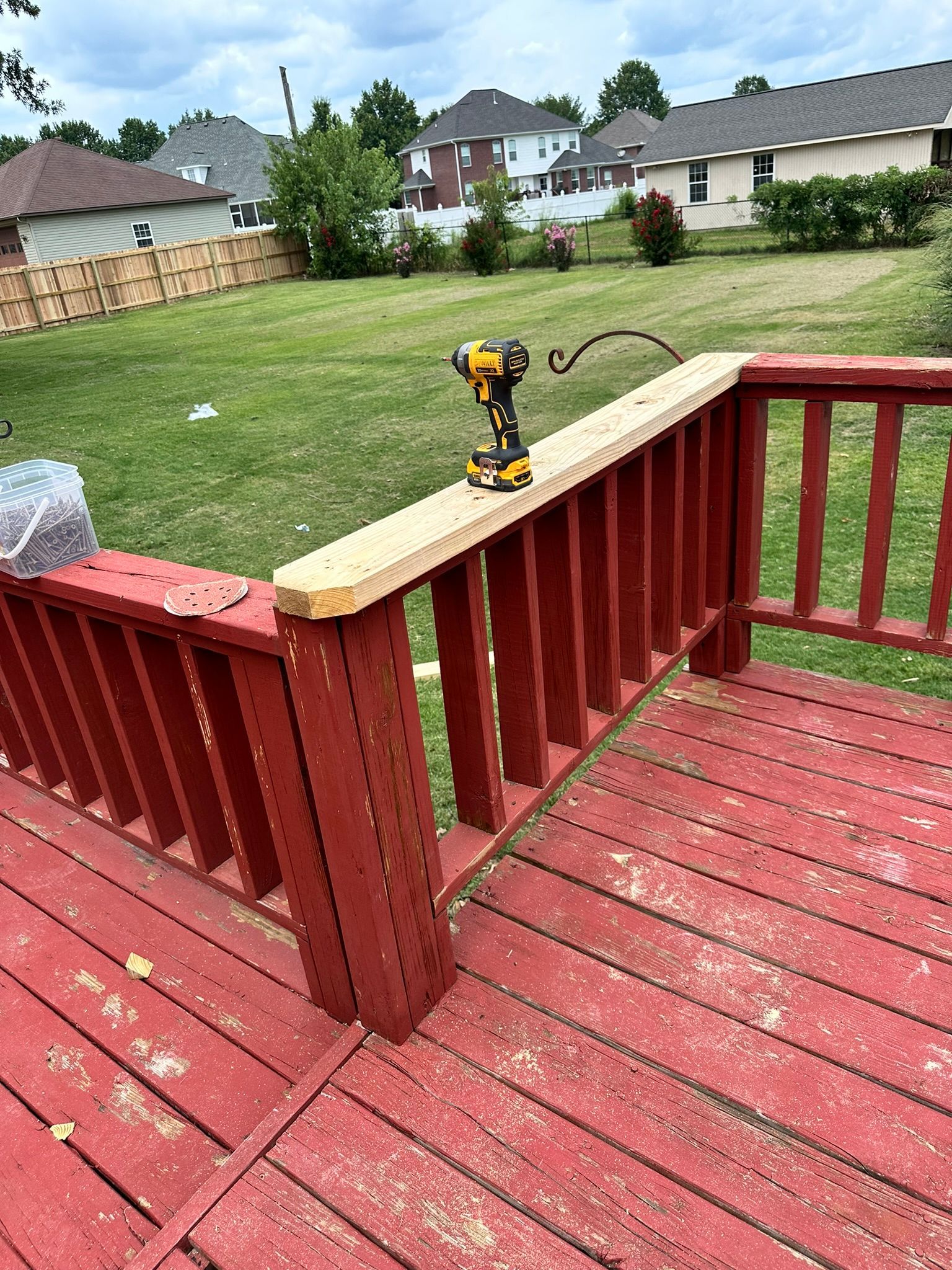 Deck railing under construction. A drill rests on a new wooden beam, over red painted deck and railing. Green lawn in the background.