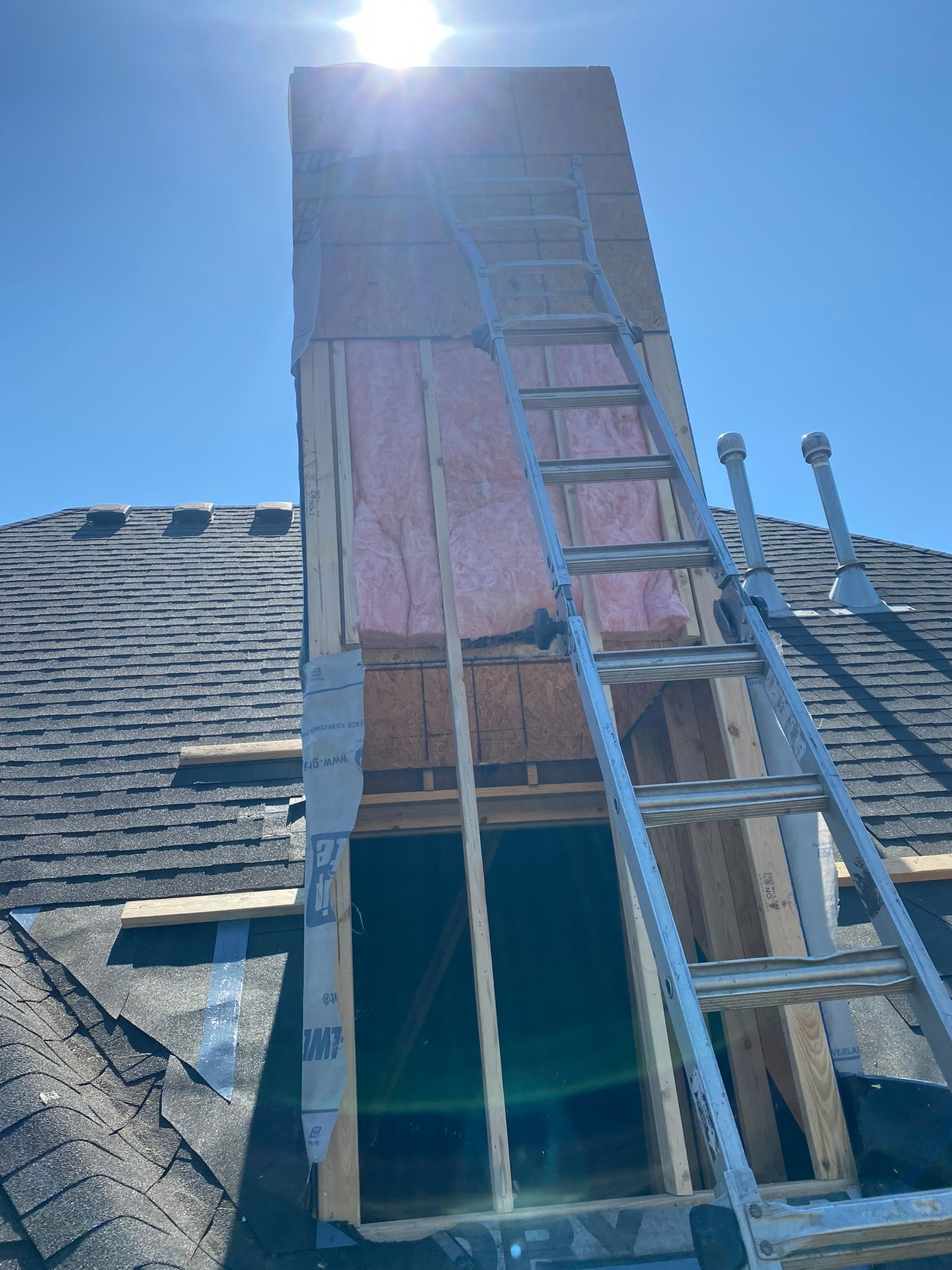 Chimney under construction on a rooftop with a ladder. Pink insulation is visible. Bright sun.