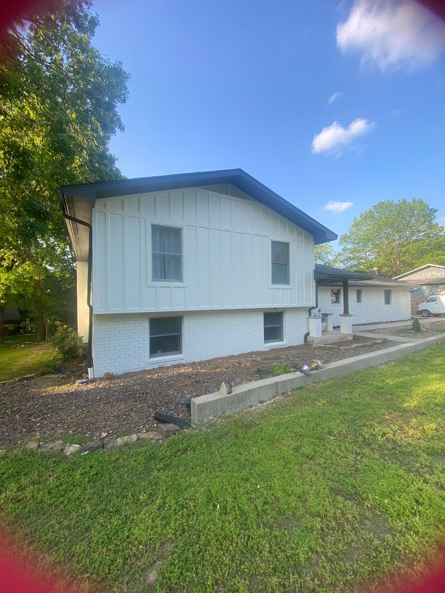 White two-story house with black roof, small windows, and landscaping in front, on a sunny day.