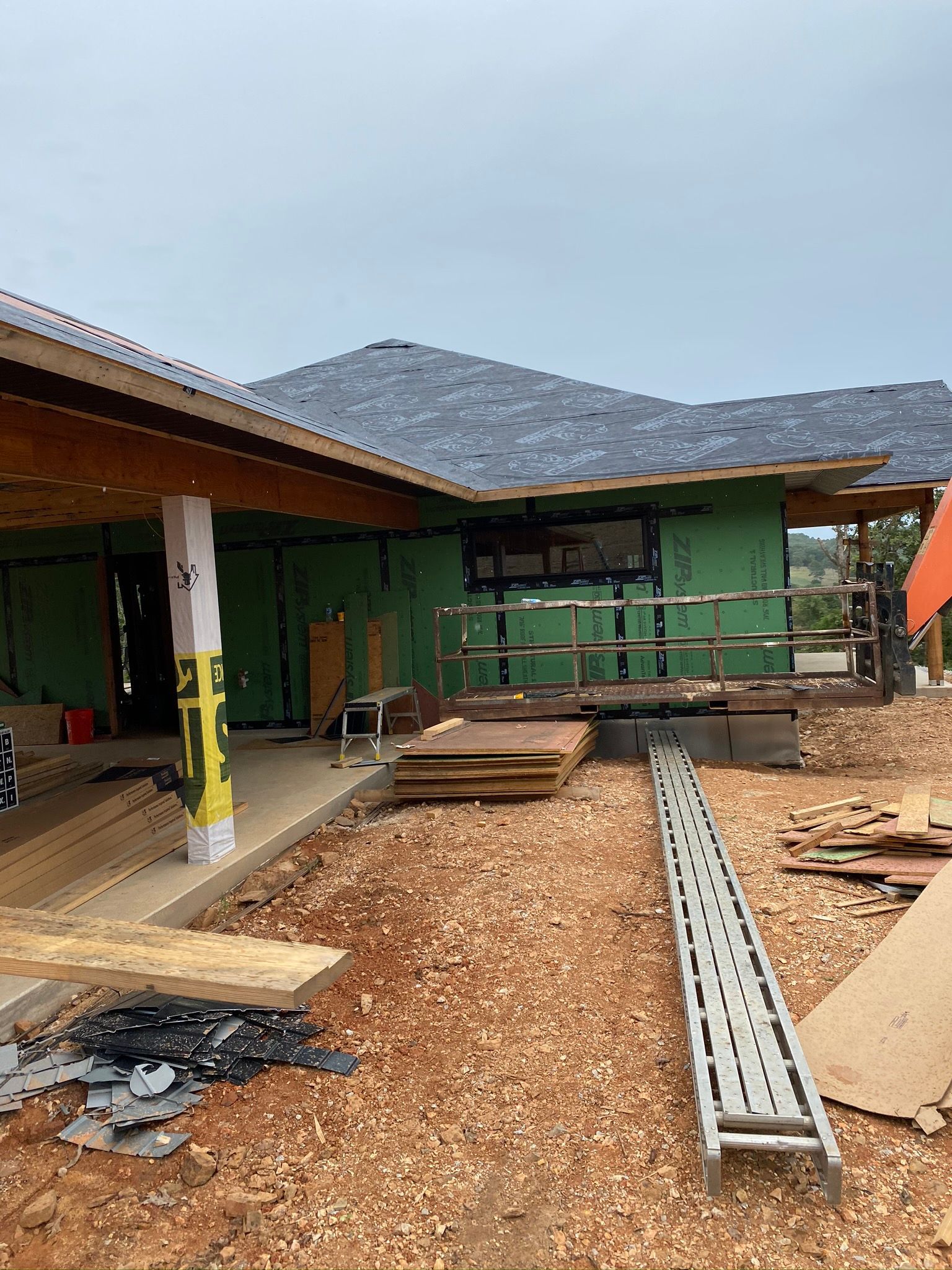 Construction of a house: wooden frame, green sheathing, dirt ground, ladder, and a large mountain in the background.