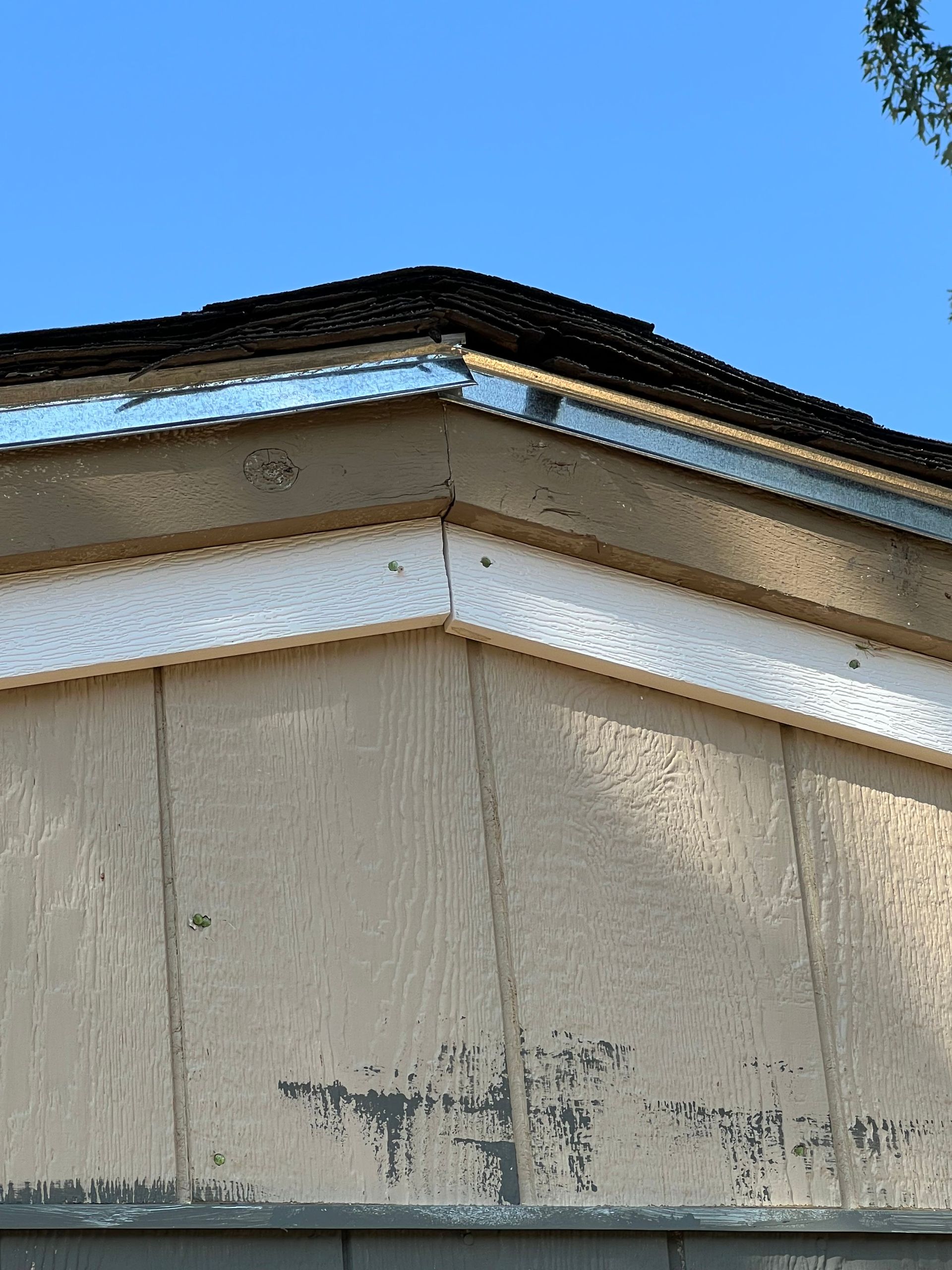 Close-up of a building's corner with weathered siding, brown trim, and a dark roof against a clear blue sky.