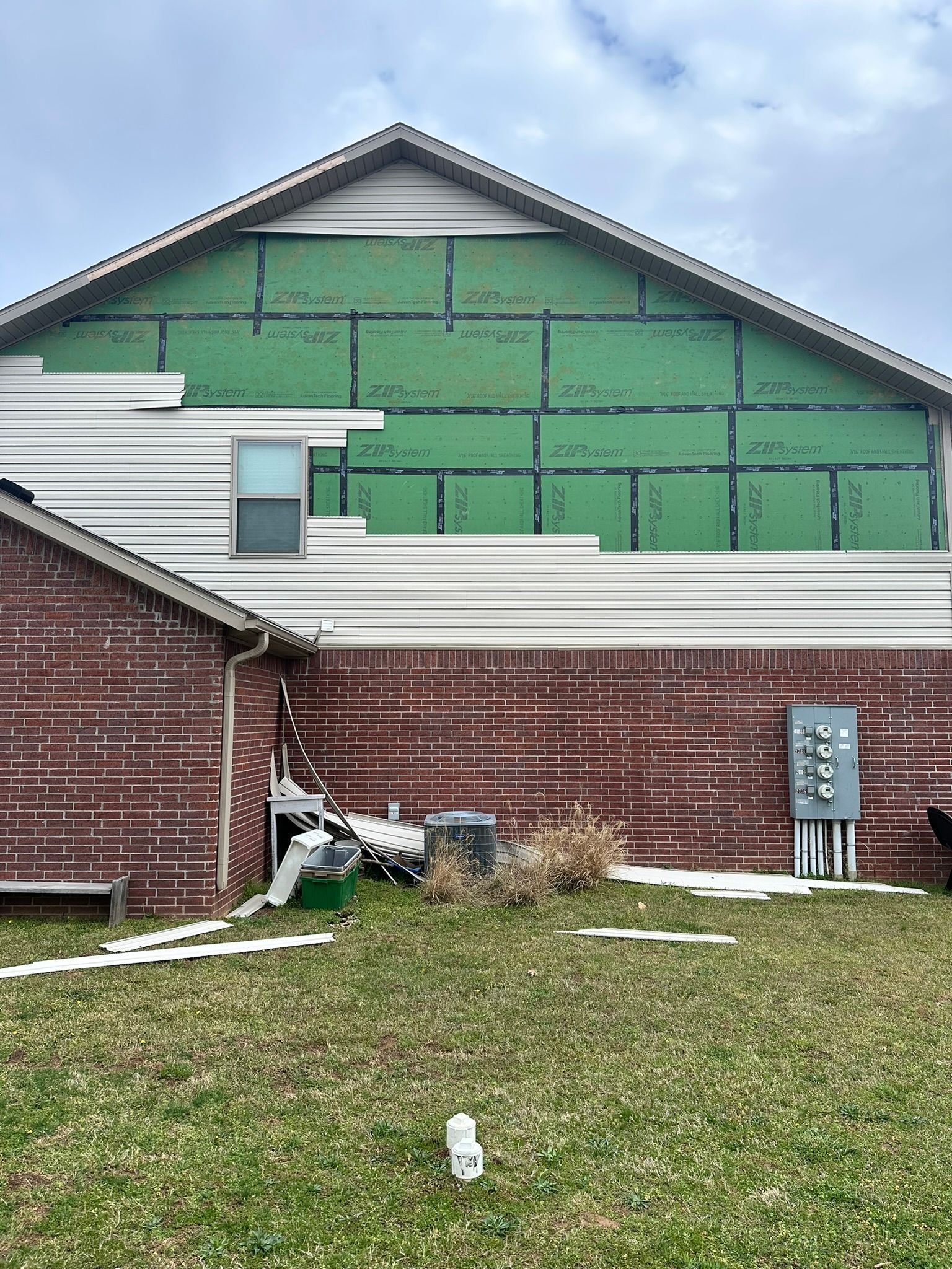 House exterior: brick lower level, siding above; green sheathing visible. Electrical box, grass.