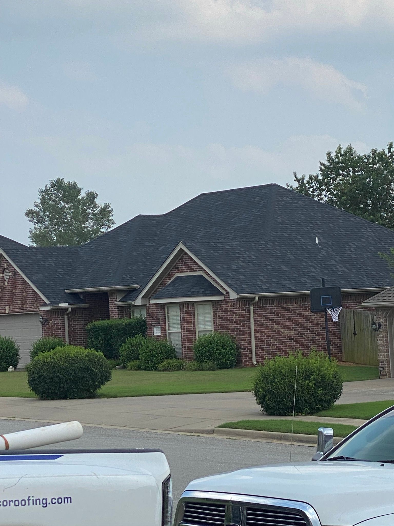 Brick house with black shingled roof, green bushes, and two parked trucks in front.